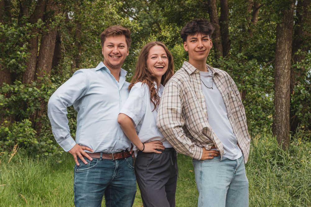Three smiling young adults posing together outdoors in a lush green forest setting on a sunny day.