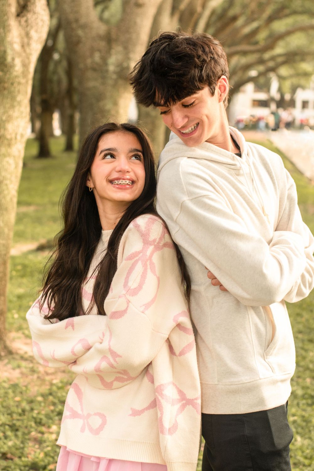Young couple in matching light sweaters share a tender moment under tree-lined path in park.