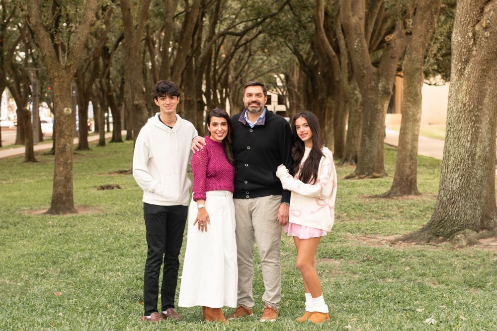 Family of four posing together on grass with tree-lined path in background.