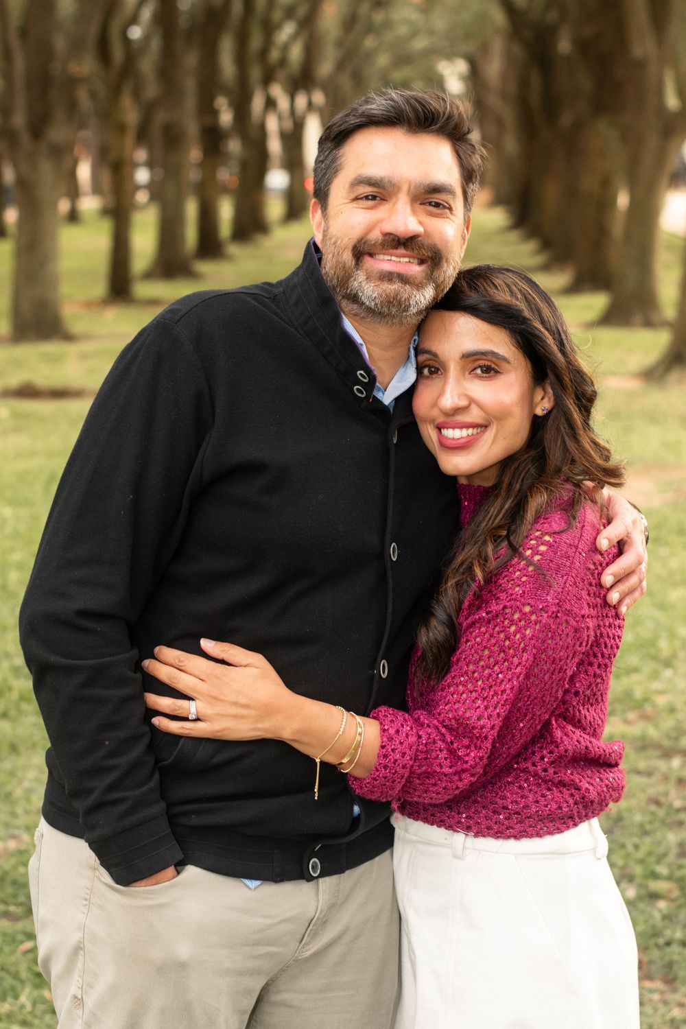 Couple embracing outdoors wearing dark cardigan and pink sweater against tree backdrop.