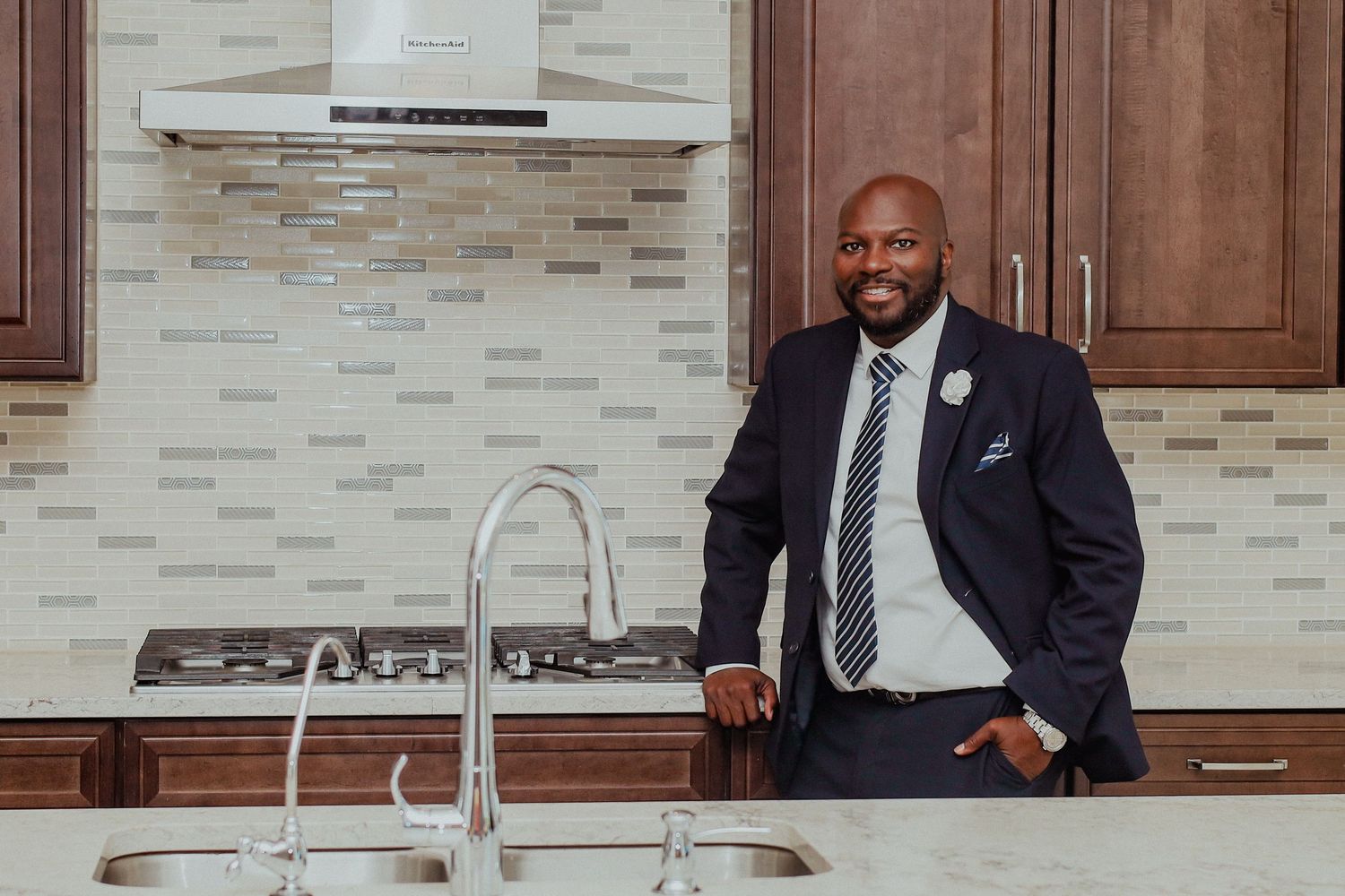 Professional man in suit smiling in modern kitchen with stainless steel appliances and mosaic backsplash.