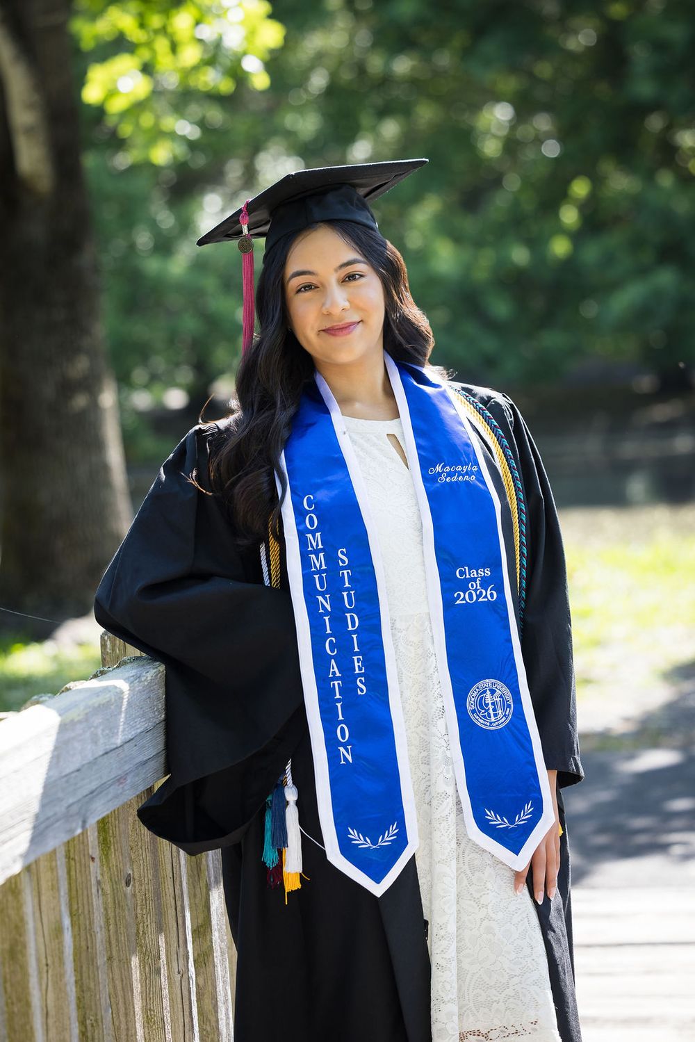 Graduate in black cap and gown with Communication Studies Class of 2026 blue stole posing outdoors.