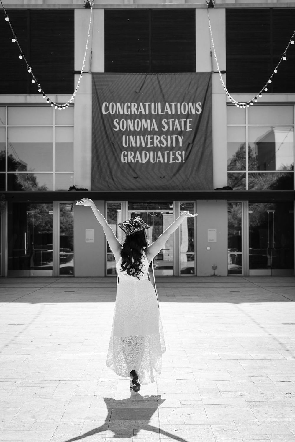 Graduate celebrates arms raised before Sonoma State University Congratulations banner in black and white.