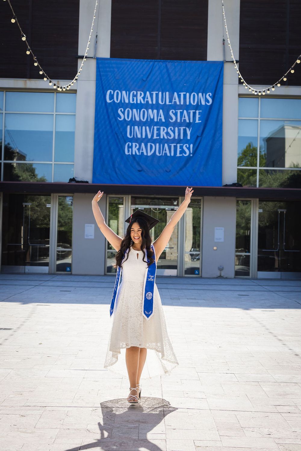 Joyful Sonoma State University graduate celebrates with arms raised in white dress and blue graduation stole.