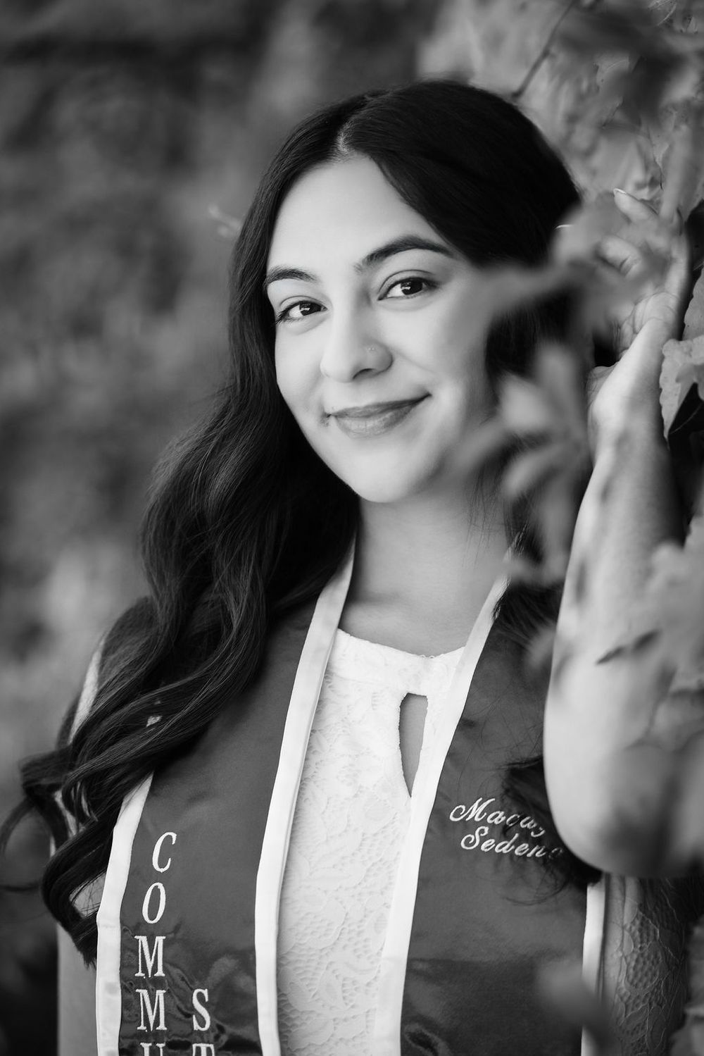 Black and white close-up portrait of smiling female graduate in sash among autumn leaves.