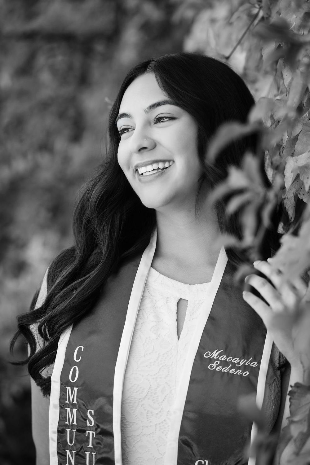 Black and white portrait of smiling young woman in graduation stole holding ivy leaves outdoors.