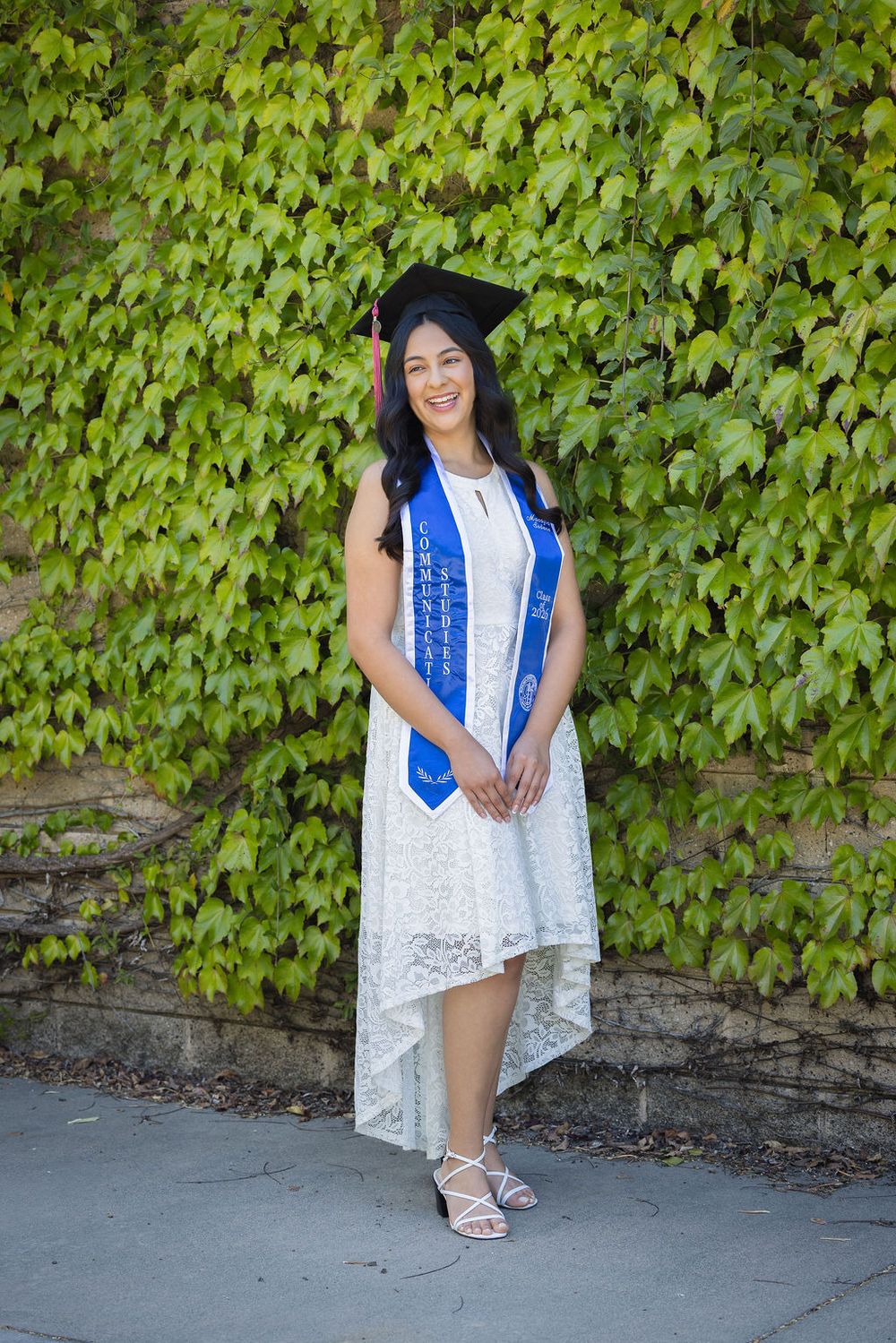 Young woman in white lace dress and graduation cap with blue honor stole poses outdoors.