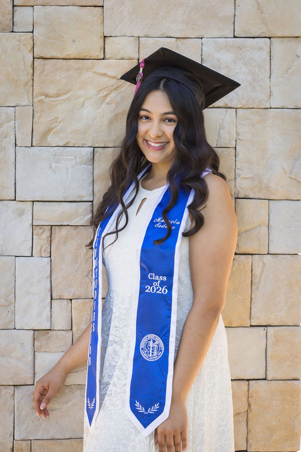 Smiling female graduate in cap and blue Class of 2026 sash poses against stone wall.