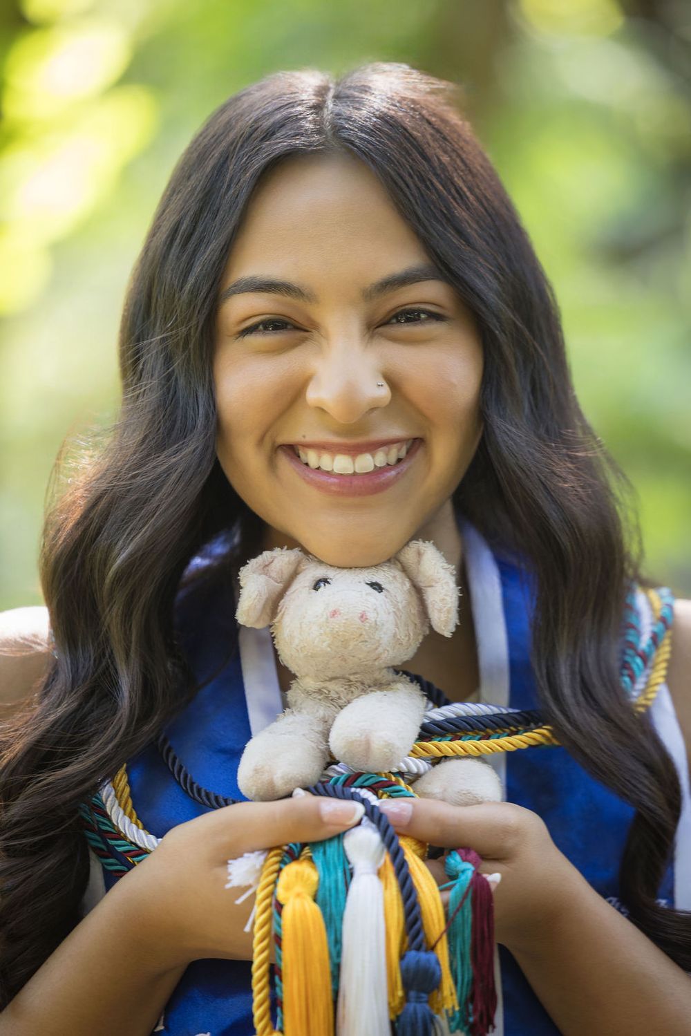Smiling young woman in graduation regalia holds a small stuffed animal with honor cords.