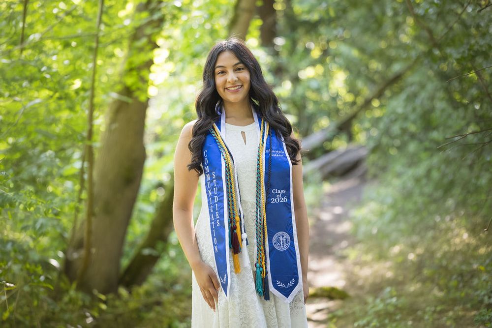 Sonoma State University graduate smiles in white dress and blue stole surrounded by lush green forest path.