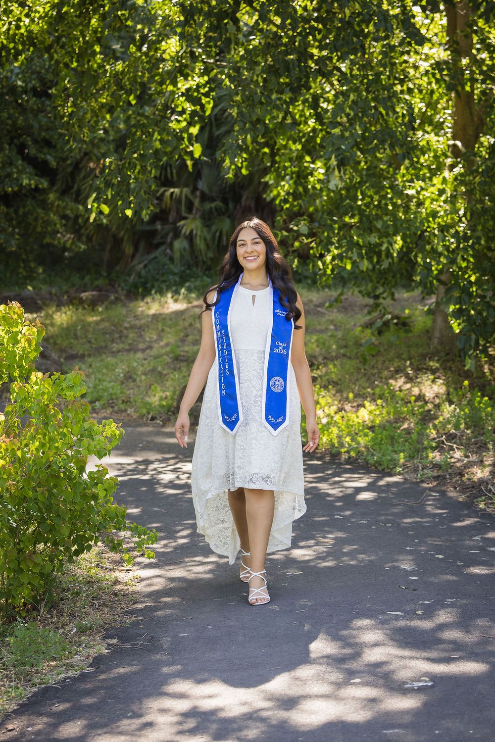 Smiling graduate in white dress and blue graduation stole walks along a tree-lined path outdoors.
