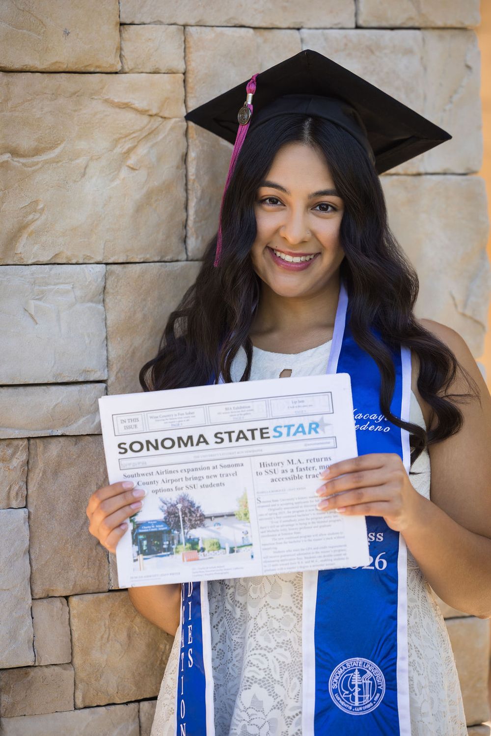 Smiling graduate in cap and blue stole holding Sonoma State Star newspaper against stone wall.