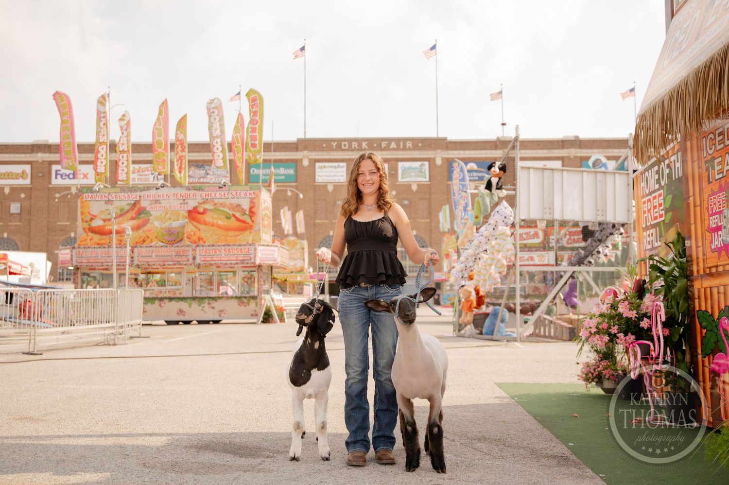 A young woman leads two goats at a county fair, wearing a black top and jeans, with colorful fair booths in the background.