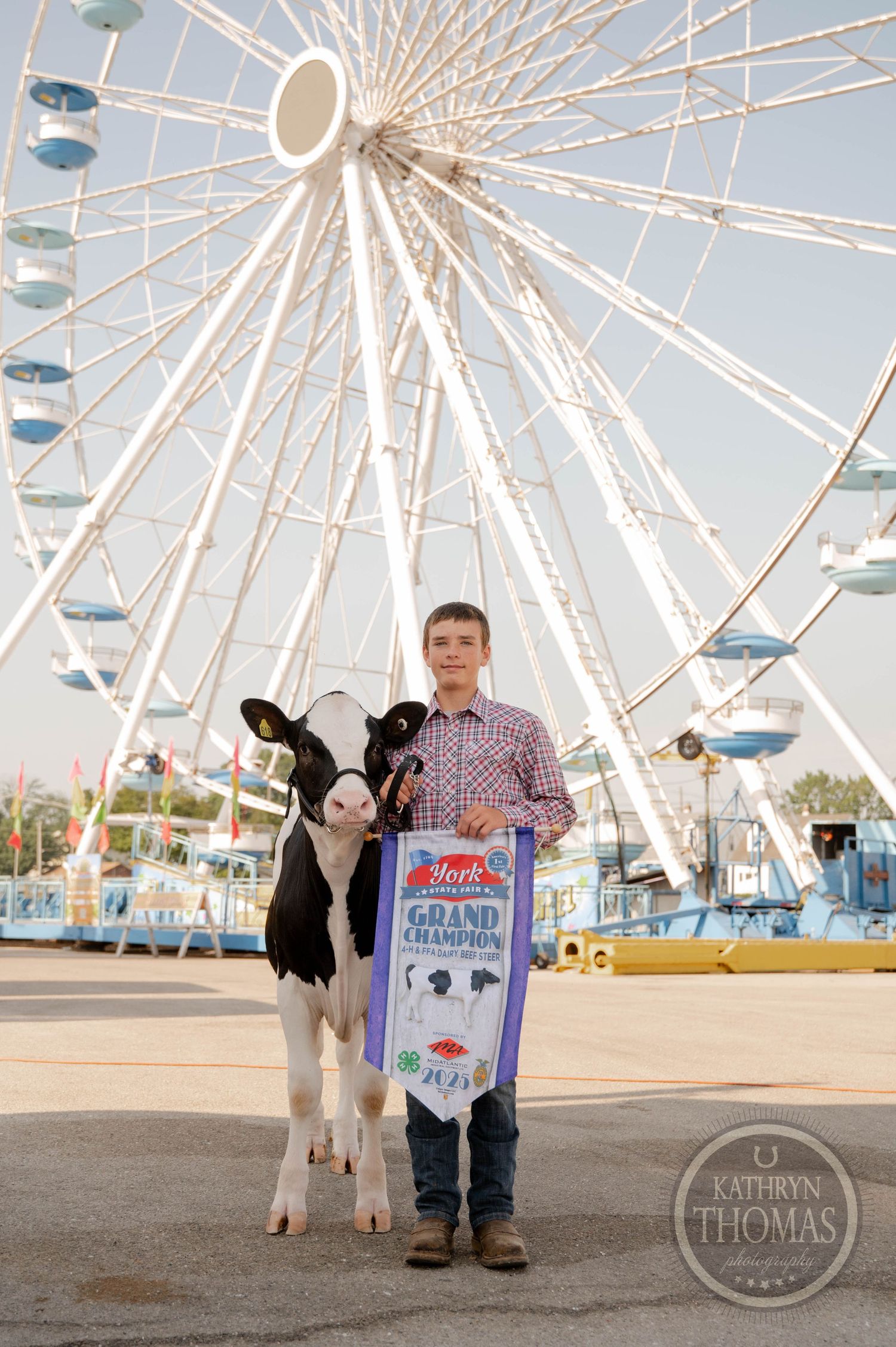 Teen boy proudly holds Grand Champion banner with Holstein calf at York County Fair, with colorful carnival booths in background.