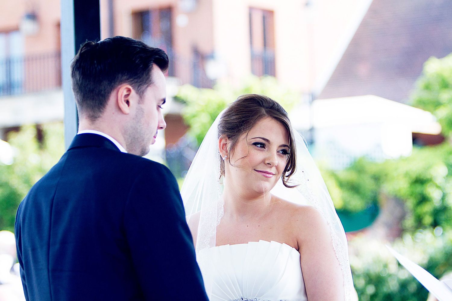 Bride in white gown and veil smiling at groom in navy suit during outdoor wedding ceremony.