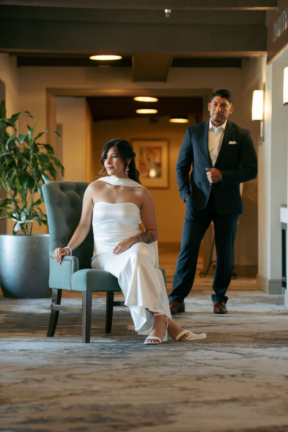 Bride in white gown seated in teal chair as groom in navy suit approaches in elegant hotel lobby.