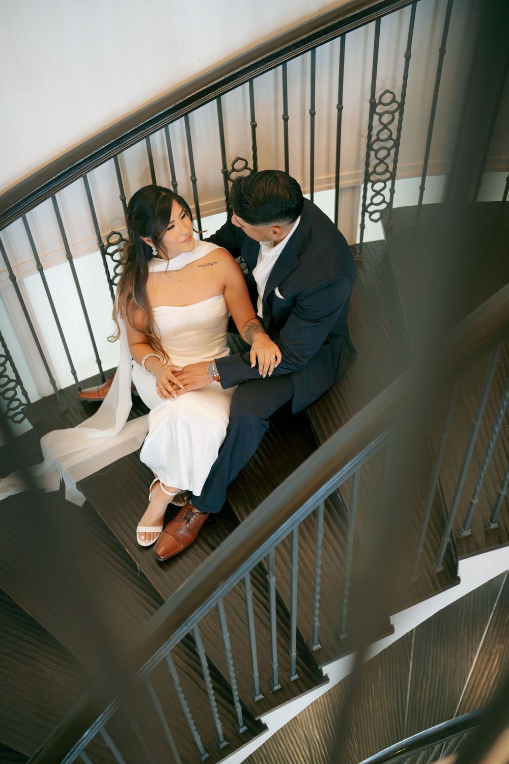 Couple in wedding attire sitting intimately on elegant spiral staircase, bride in white dress, groom in navy suit.