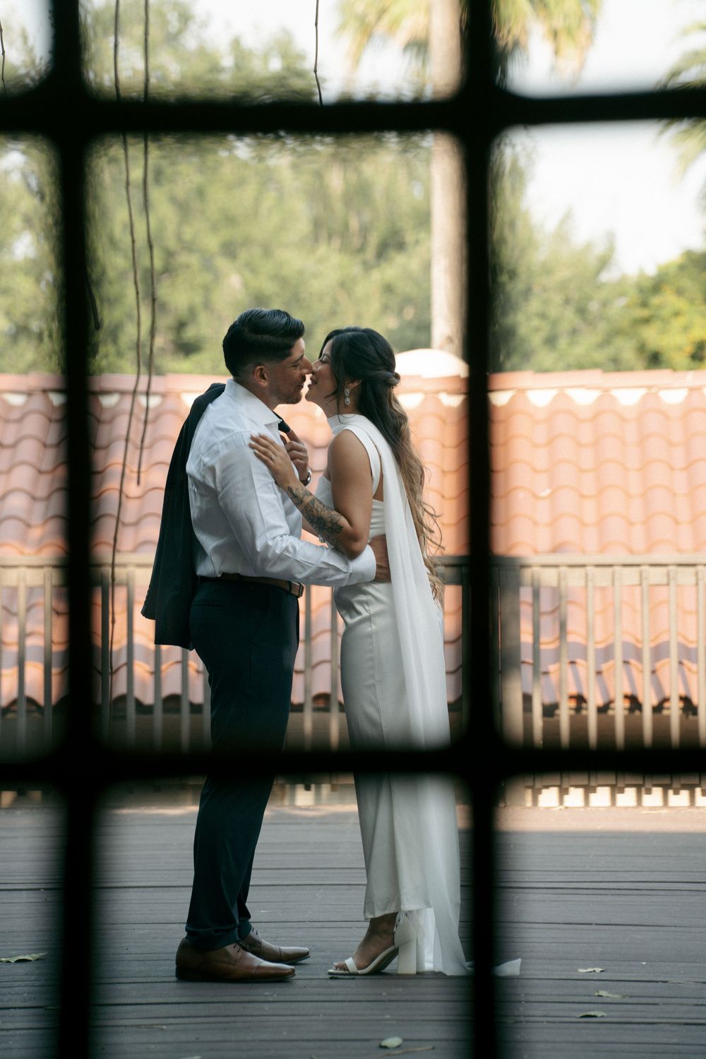 Bride and groom share a romantic moment on a balcony, viewed through a window frame, with terracotta rooftops behind.