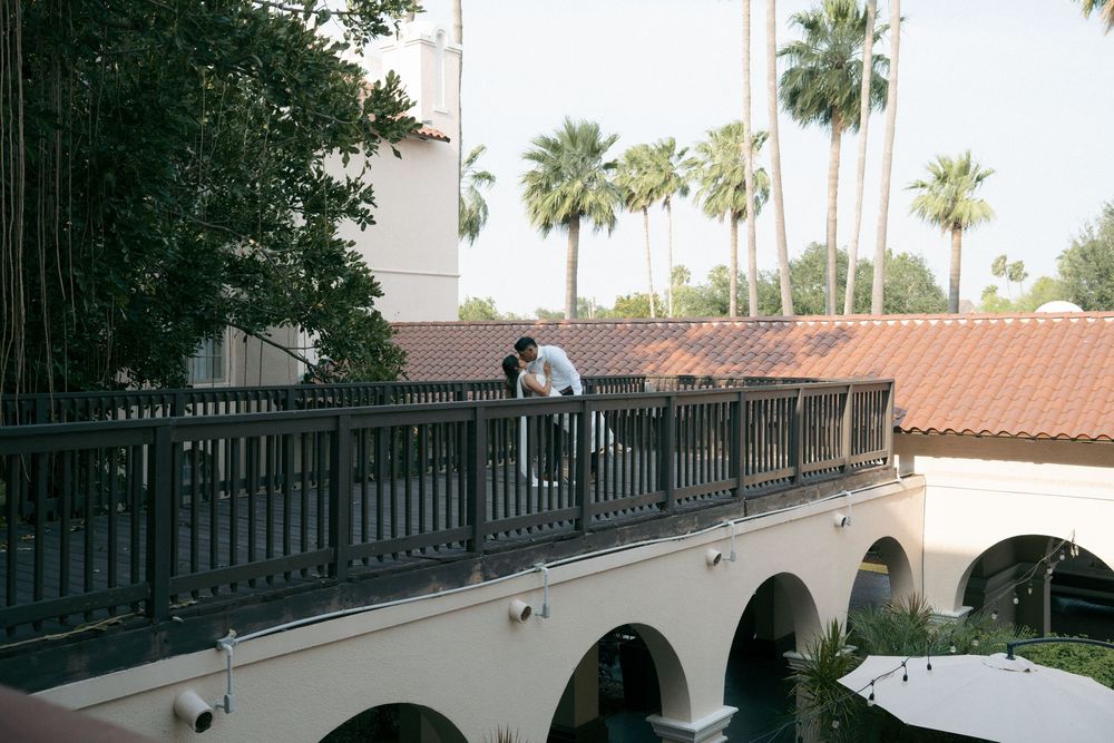A couple shares a romantic moment on a balcony overlooking arched walkways and palm trees at a resort.