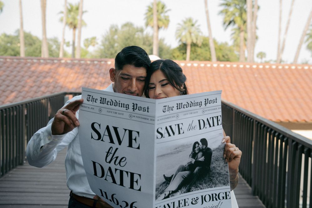 Couple holding a Save the Date newspaper announcement for Kaylee and Jorge wedding outdoors on a wooden bridge.
