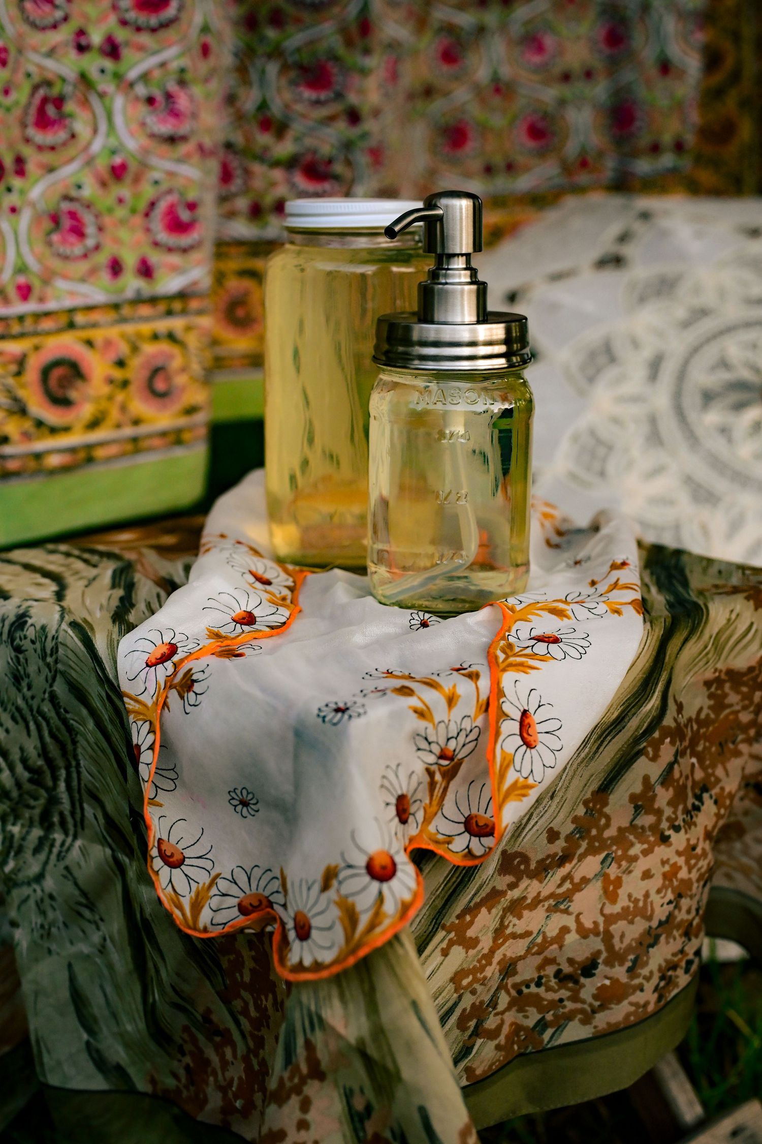 Two glass mason jar dispensers with yellow liquid on floral fabric, surrounded by colorful boho textiles.
