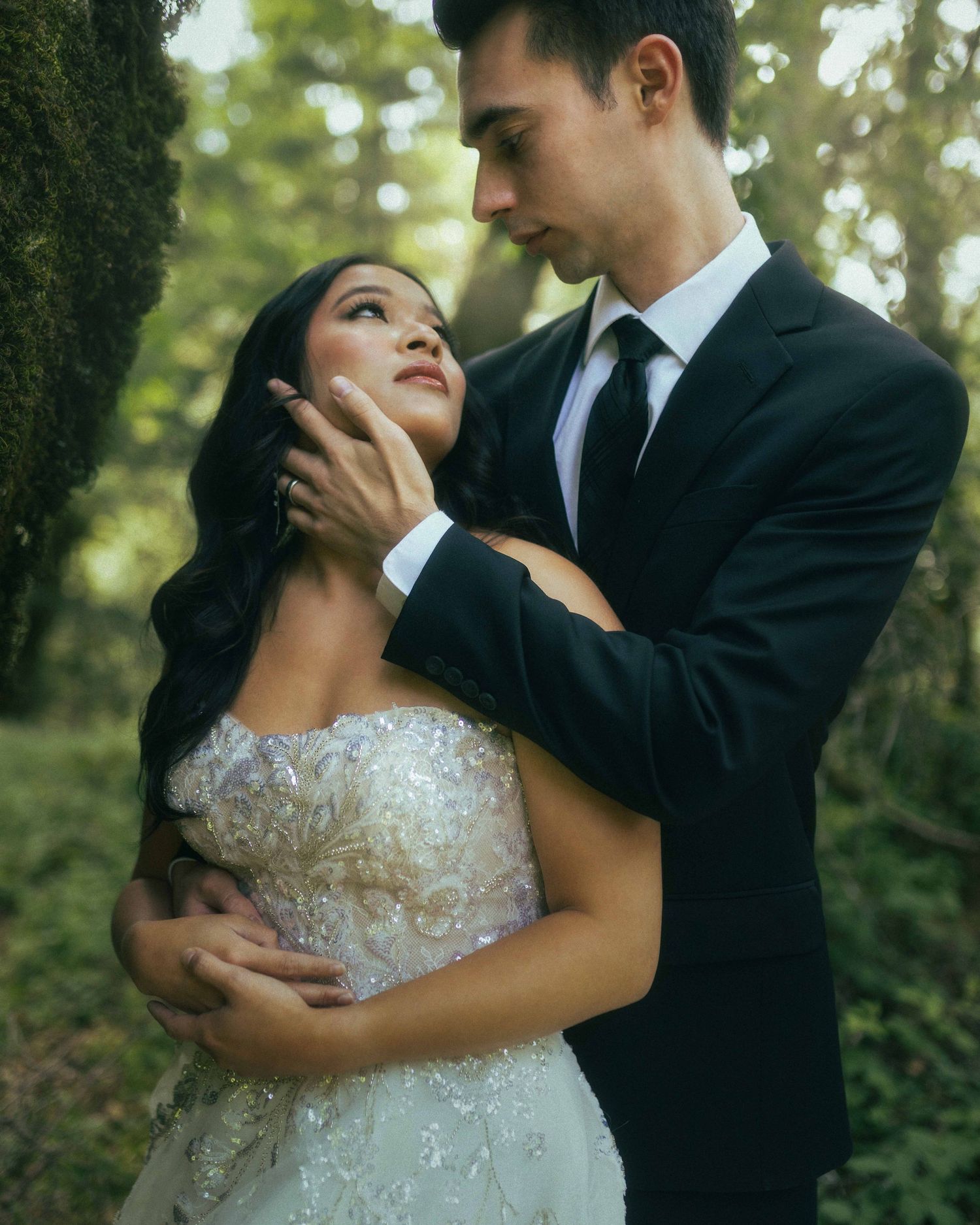 Bride and groom share a romantic embrace in a lush forest, groom in black suit gently cupping brides face.