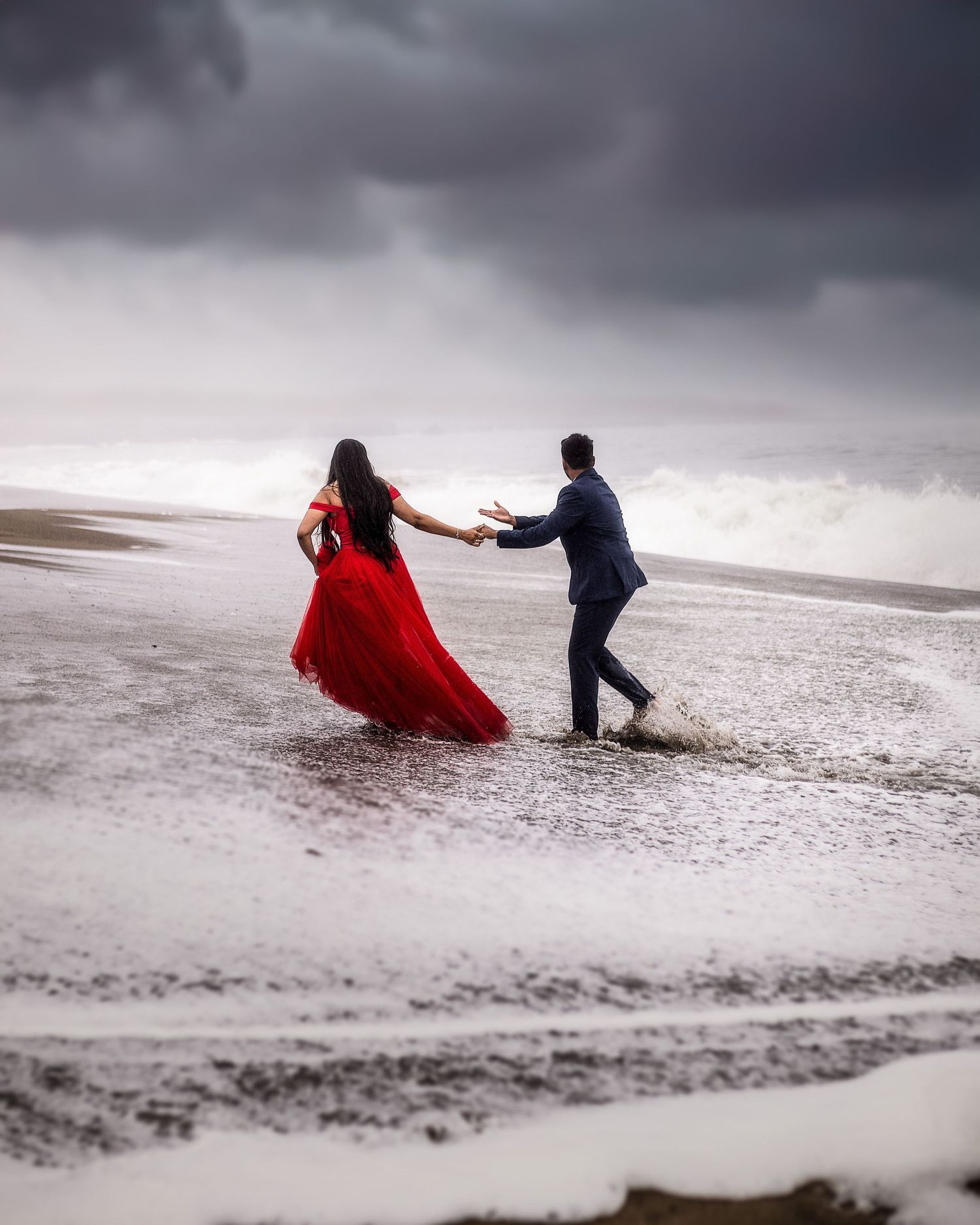 Couple dancing on stormy beach, woman in flowing red gown, man in navy suit, dramatic cloudy sky.