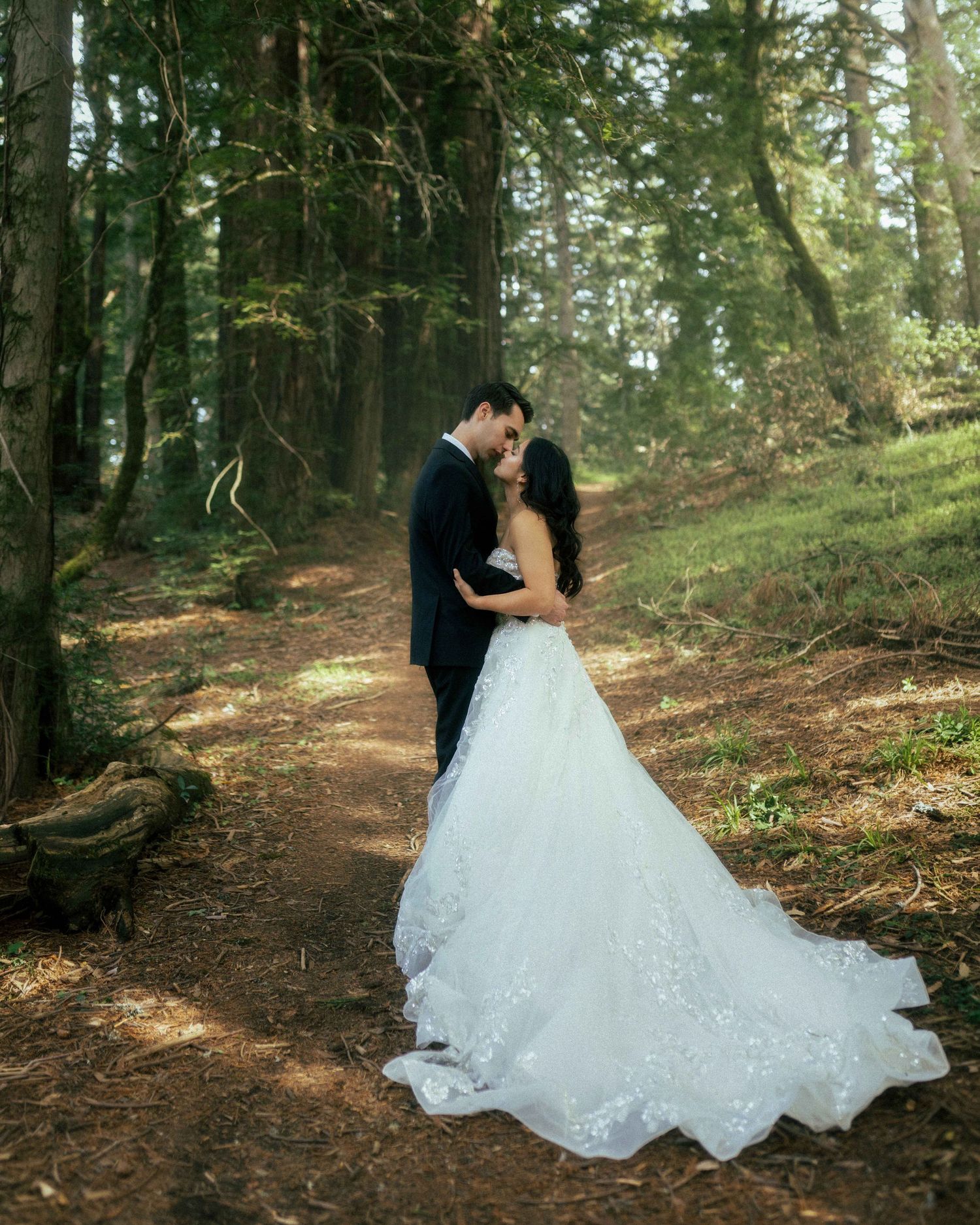 Wedding couple sharing a kiss on a sunlit forest path, bride in elegant lace gown with long train.