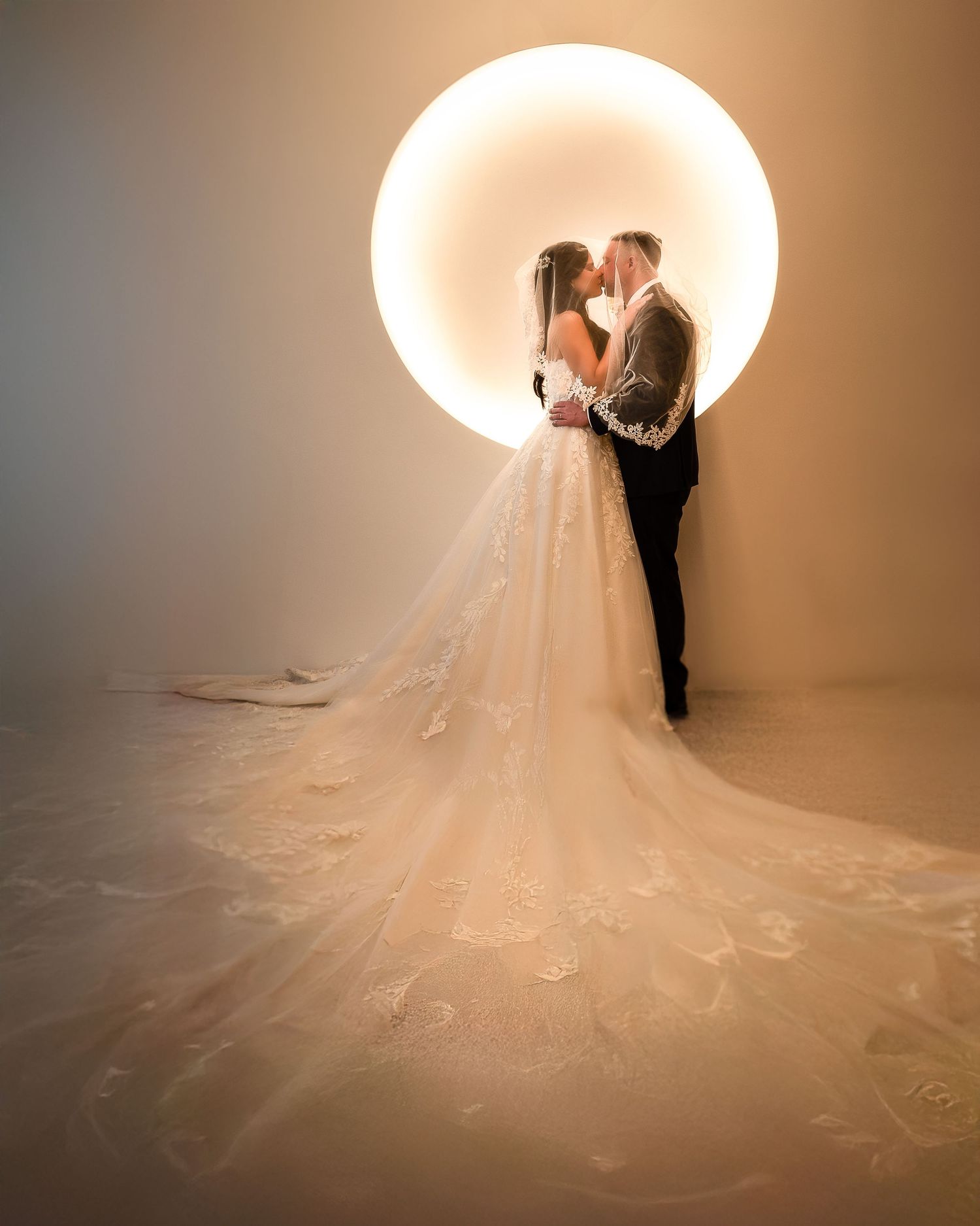 Couple shares an intimate kiss silhouetted against a glowing circular light backdrop in elegant wedding attire.