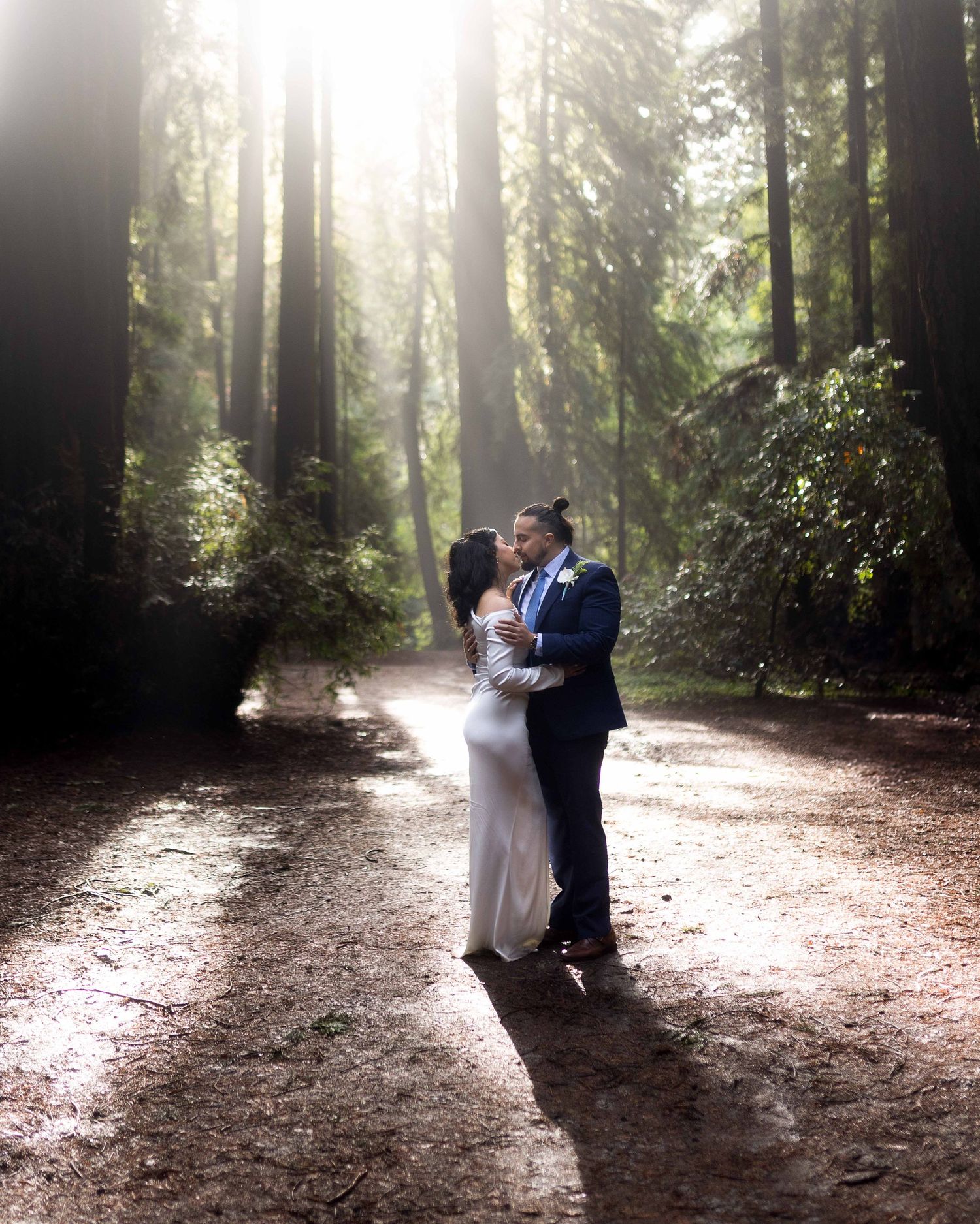 Couple embraces on a sunlit forest path surrounded by towering redwood trees in a romantic wedding photo.