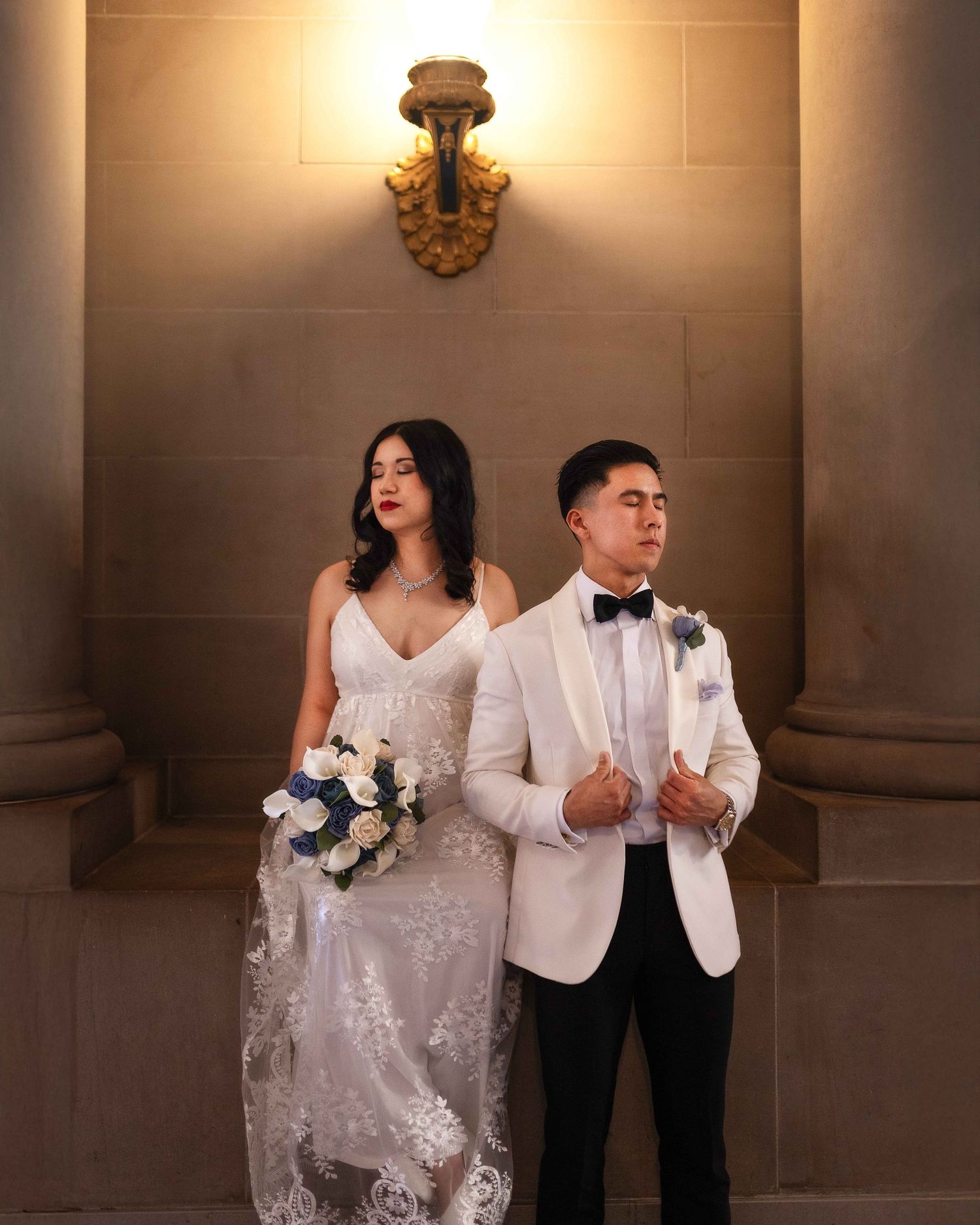 Bride and groom posing in ornate indoor venue, groom in white tuxedo and bride holding white bouquet.
