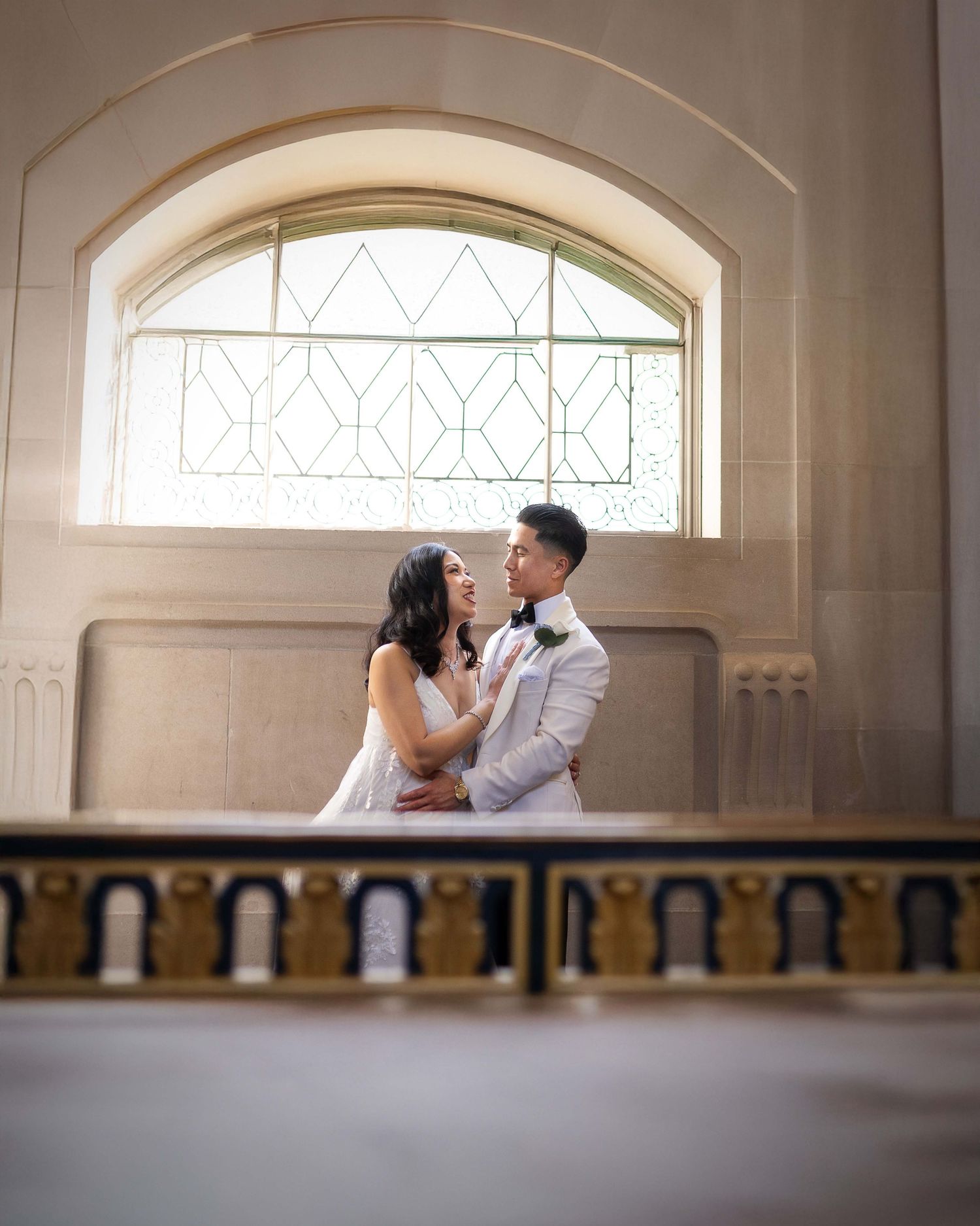 Bride and groom share a tender moment beneath an arched stained glass window in a grand historic venue.