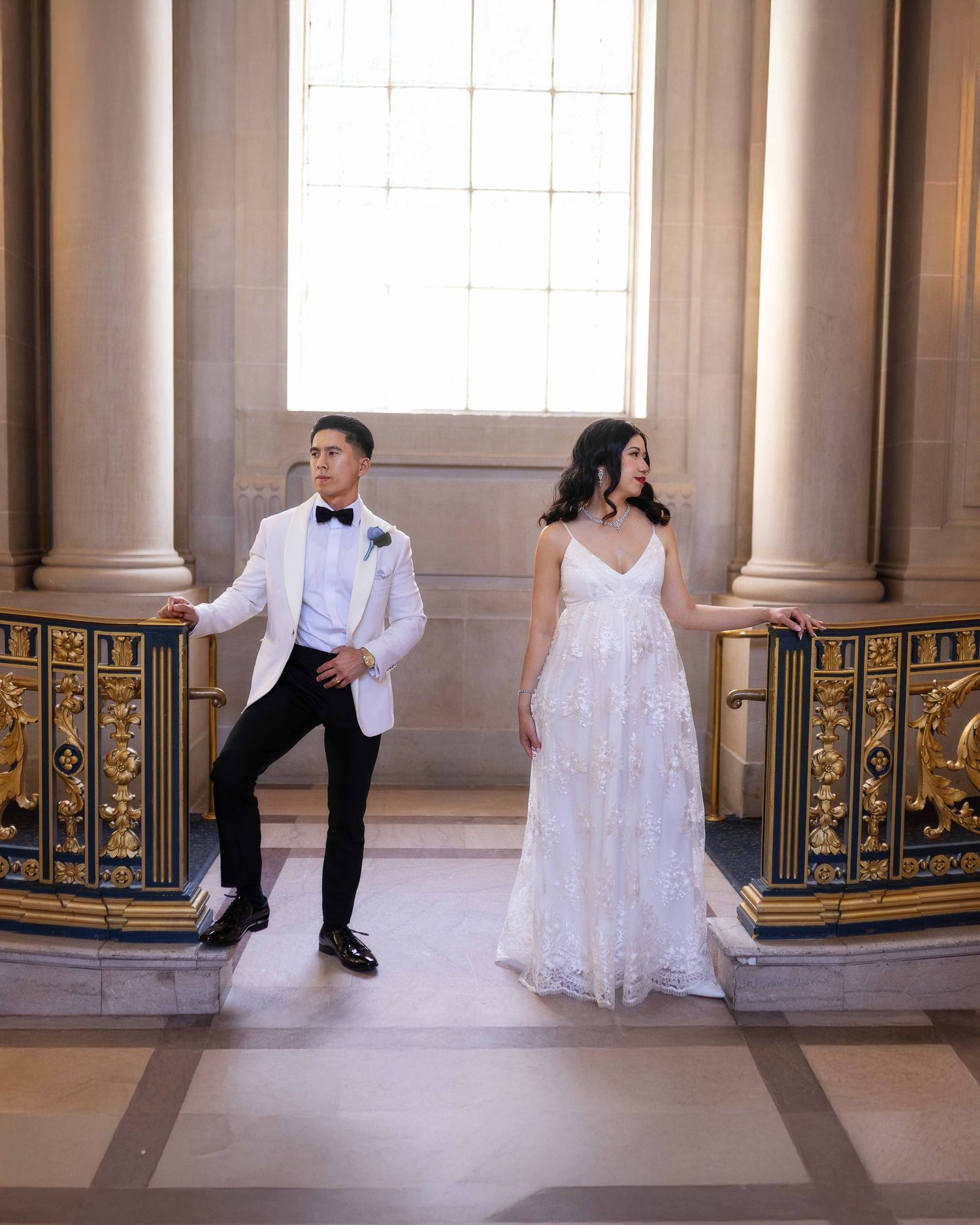 Bride in floral gown and groom in white tuxedo pose elegantly on ornate gold staircase at San Francisco City Hall.