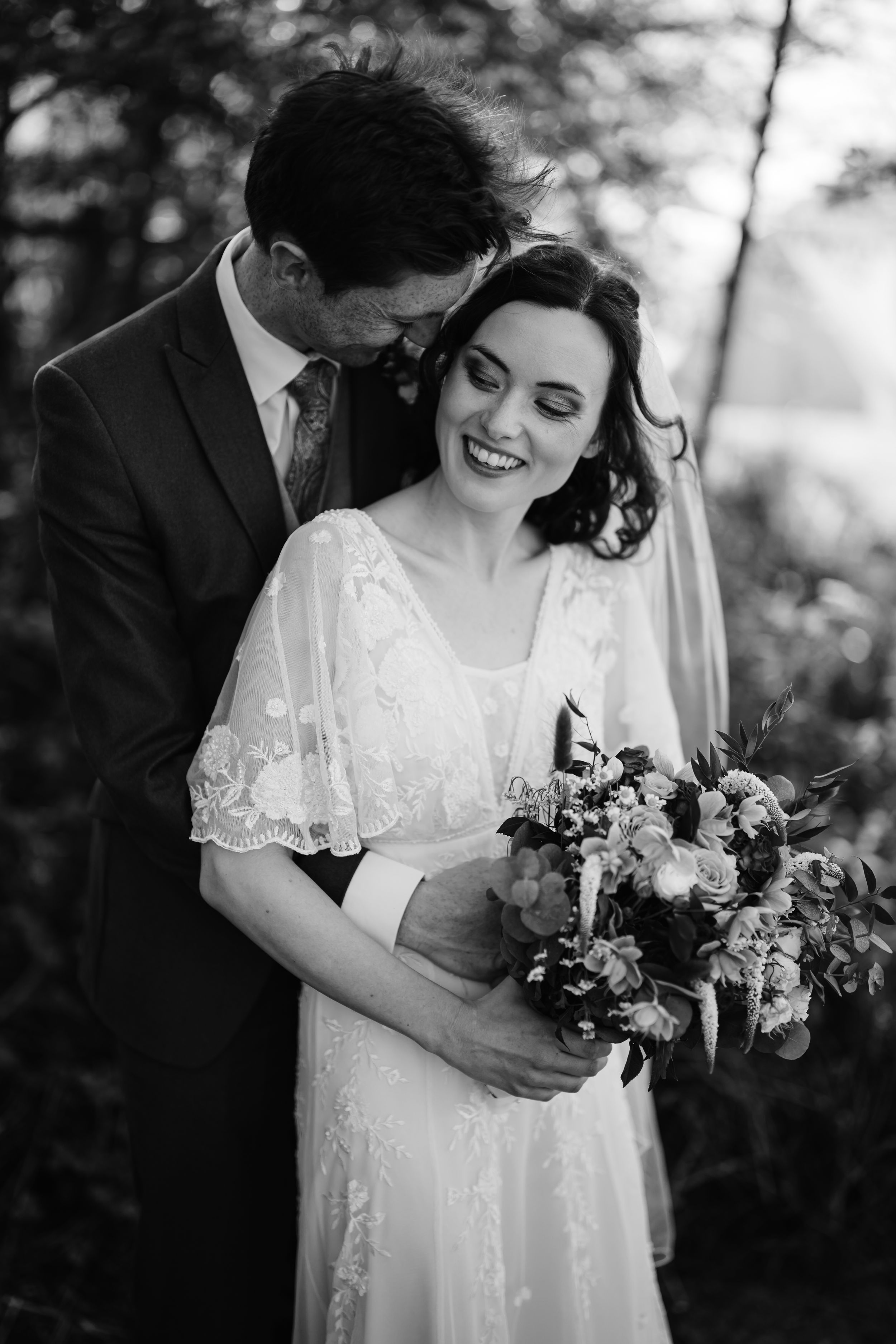 Black and white photo of smiling bride holding bouquet embraced by groom in suit outdoors.
