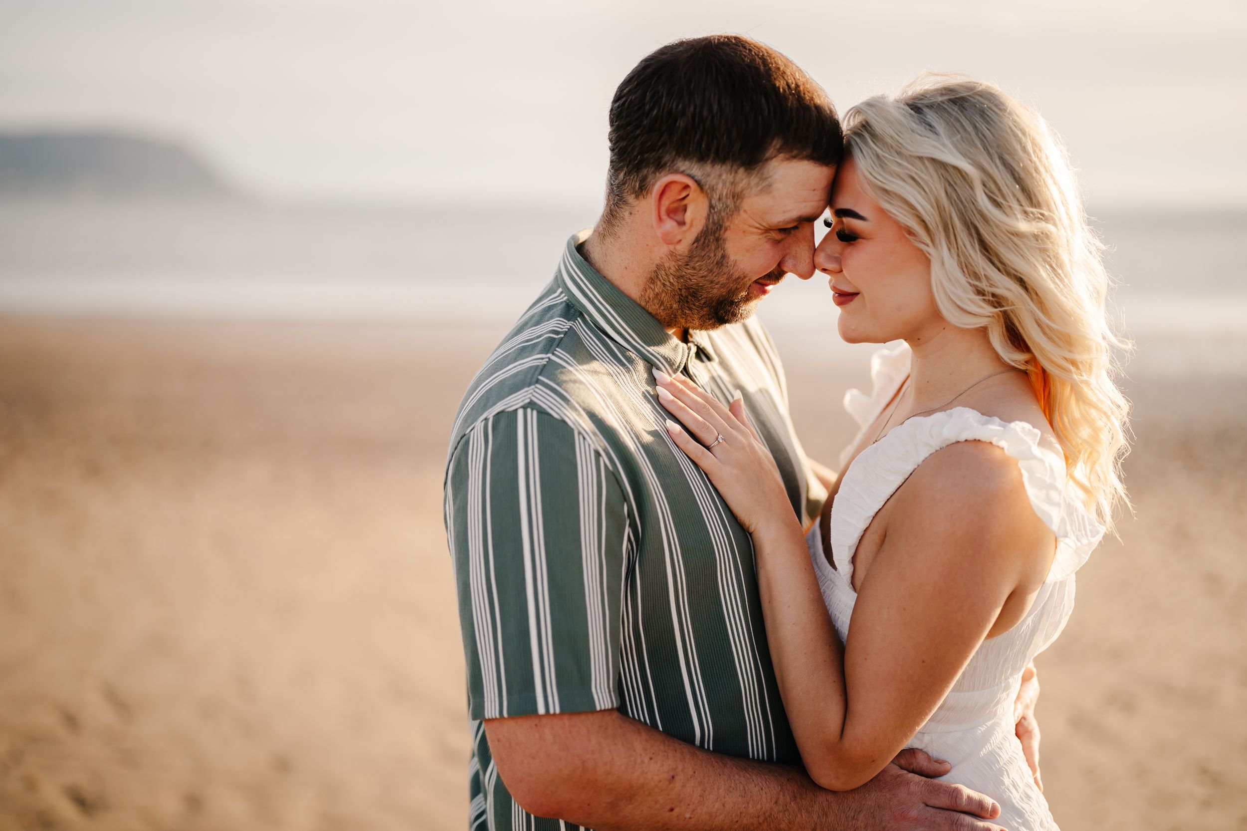 Romantic couple touching foreheads on a sandy beach at golden hour, woman in white top, man in striped shirt.