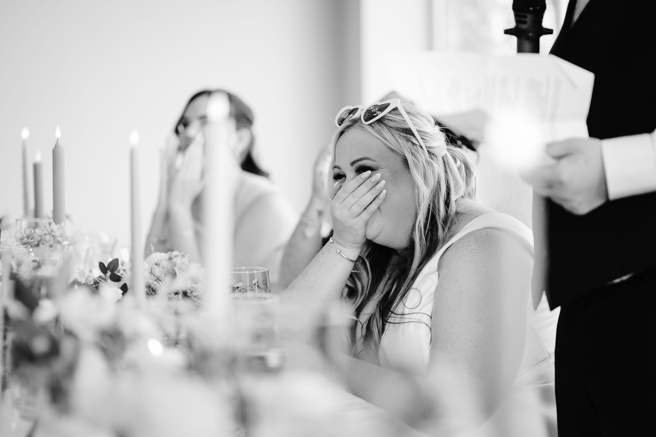 Black and white wedding reception photo of a laughing bride covering her mouth during a heartfelt speech.