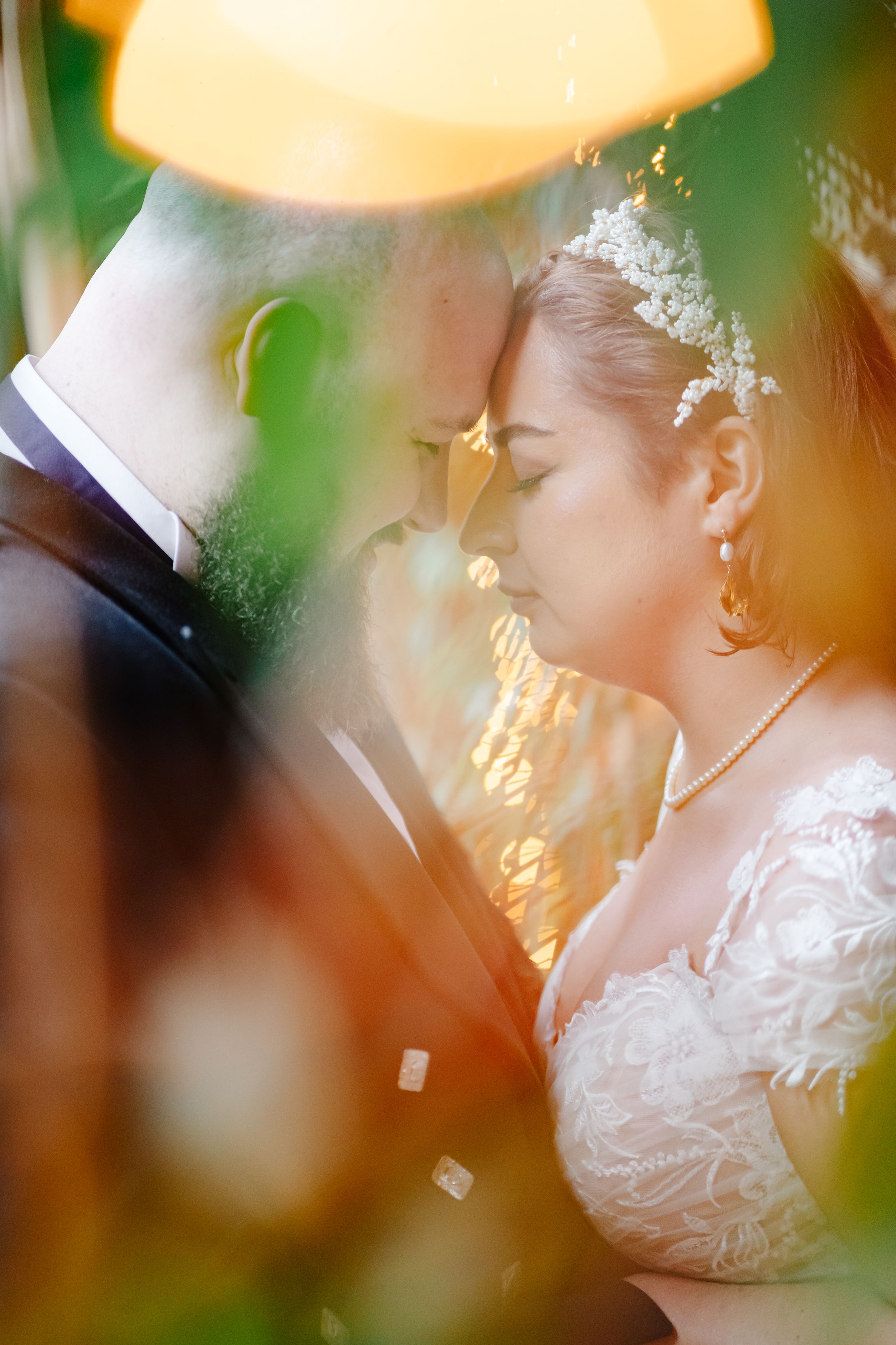 Bride and groom share an intimate forehead touch, bathed in warm golden light, surrounded by soft bokeh greenery.