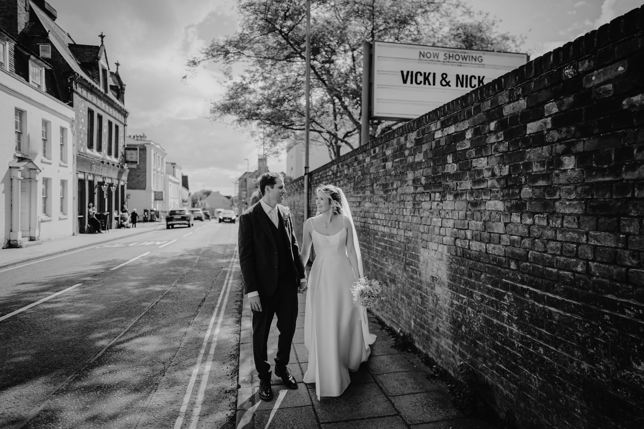 Black and white wedding photo of bride and groom walking along a brick wall street, with a Vicki and Nick sign above.