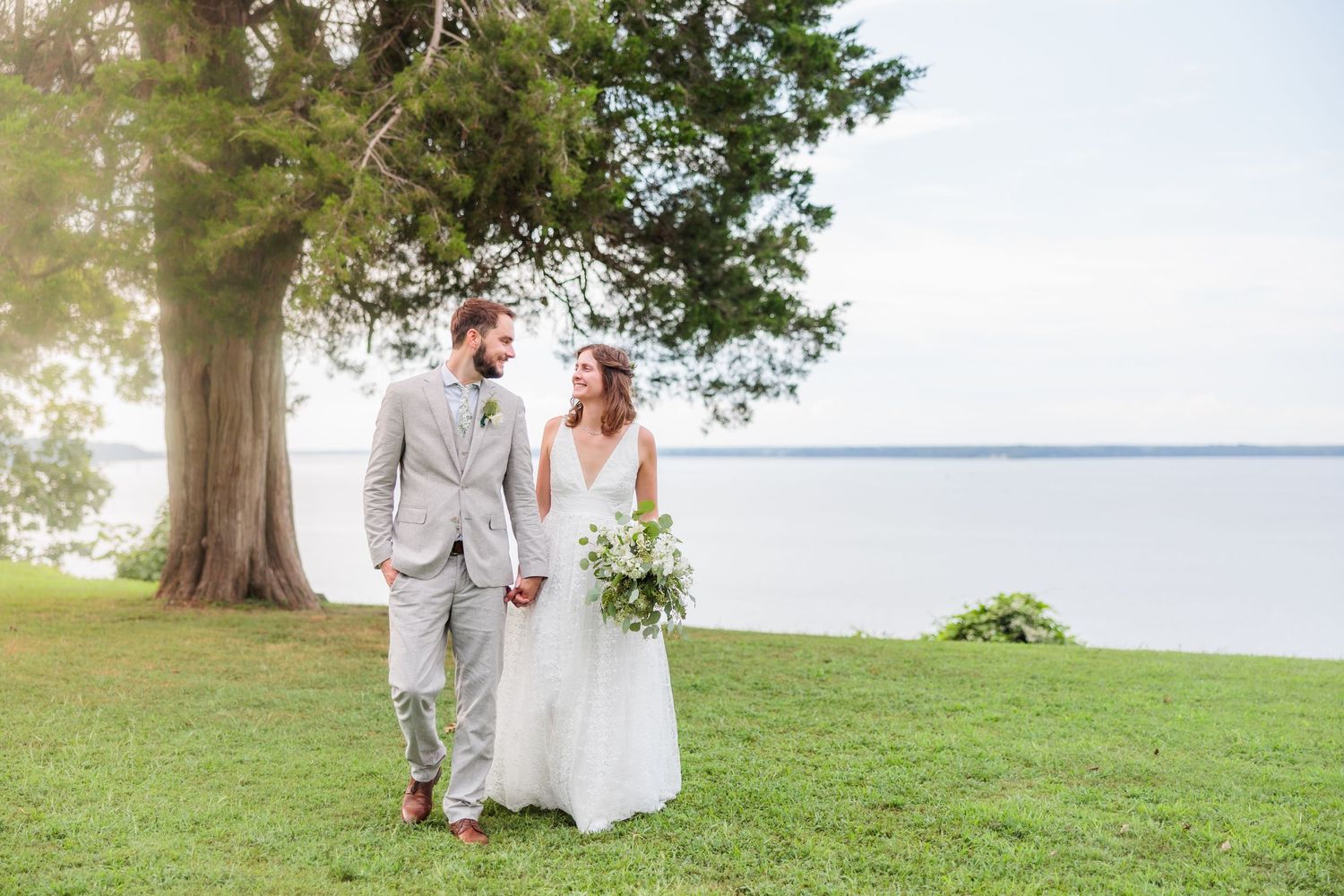 Wedding couple walking hand in hand on grassy lawn by waterfront.