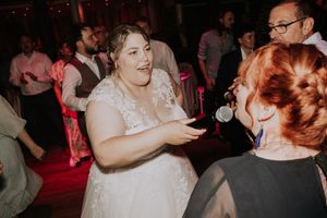 Bride in white lace gown dancing joyfully with guests at a wedding reception under warm pink lights.