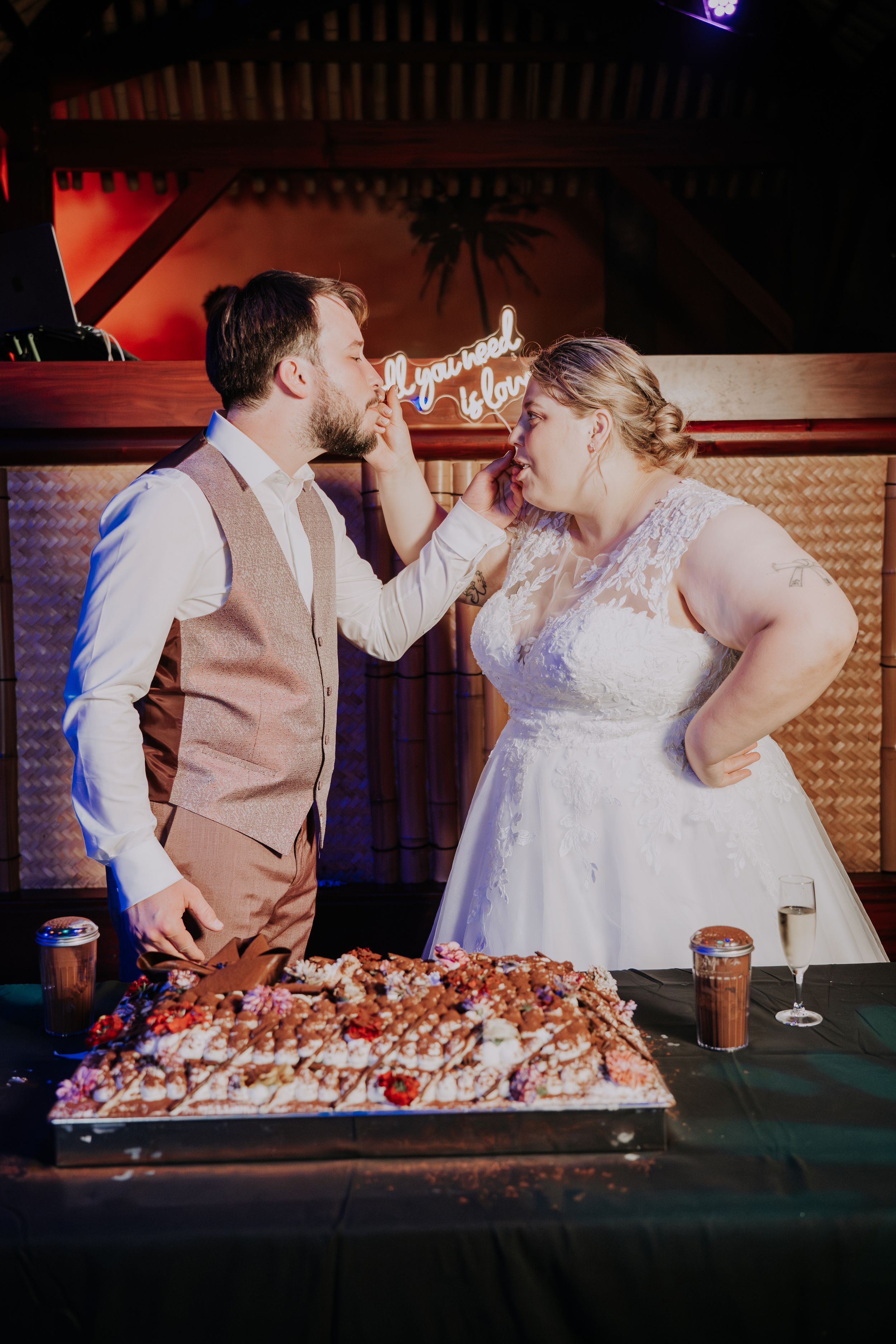 Bride and groom feeding each other cake at wedding reception with neon sign backdrop and large decorated sheet cake.