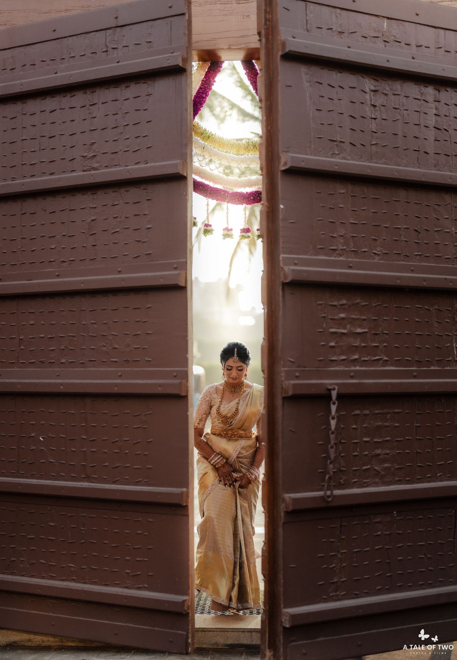 Indian bride in gold saree framed by large wooden doors at traditional wedding ceremony.