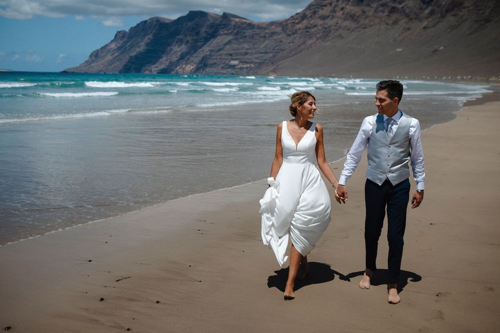 Newlywed couple walking hand in hand on a sandy beach with turquoise ocean waves and dramatic mountains behind them.