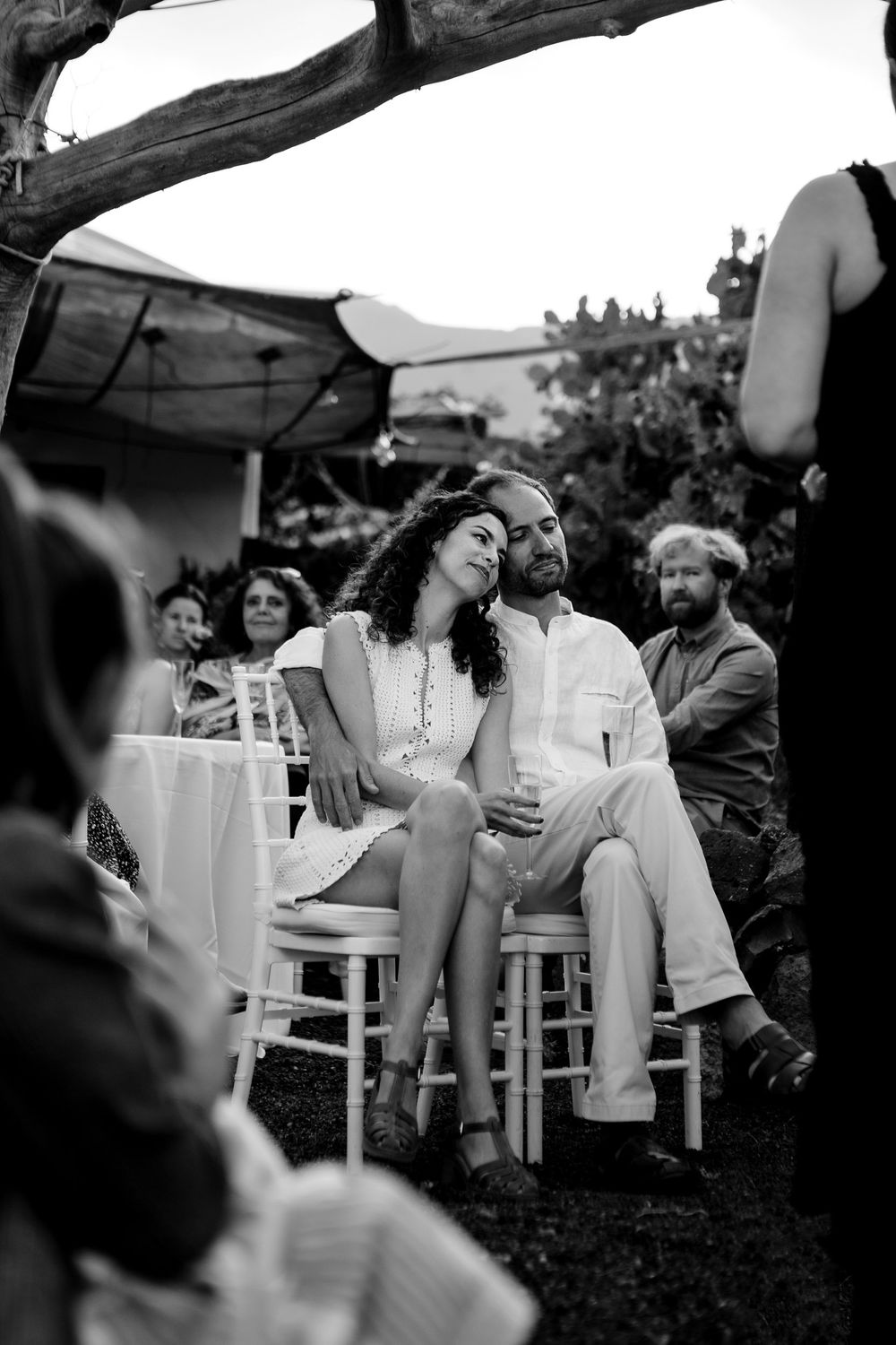 Black and white photo of a couple in white attire sitting closely together at an outdoor wedding ceremony.