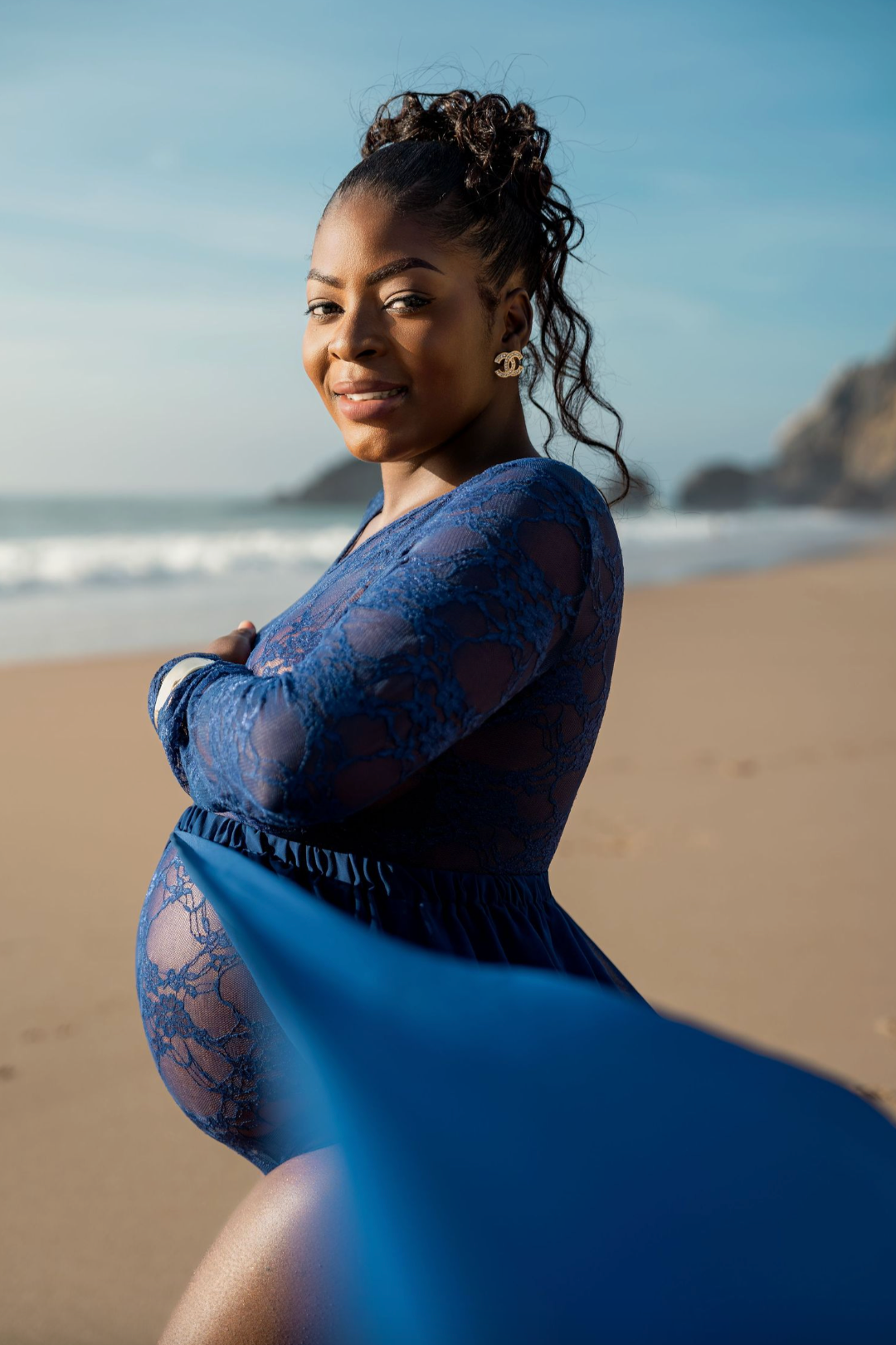Pregnant woman in blue lace dress posing gracefully on a sunny beach with ocean waves in the background.