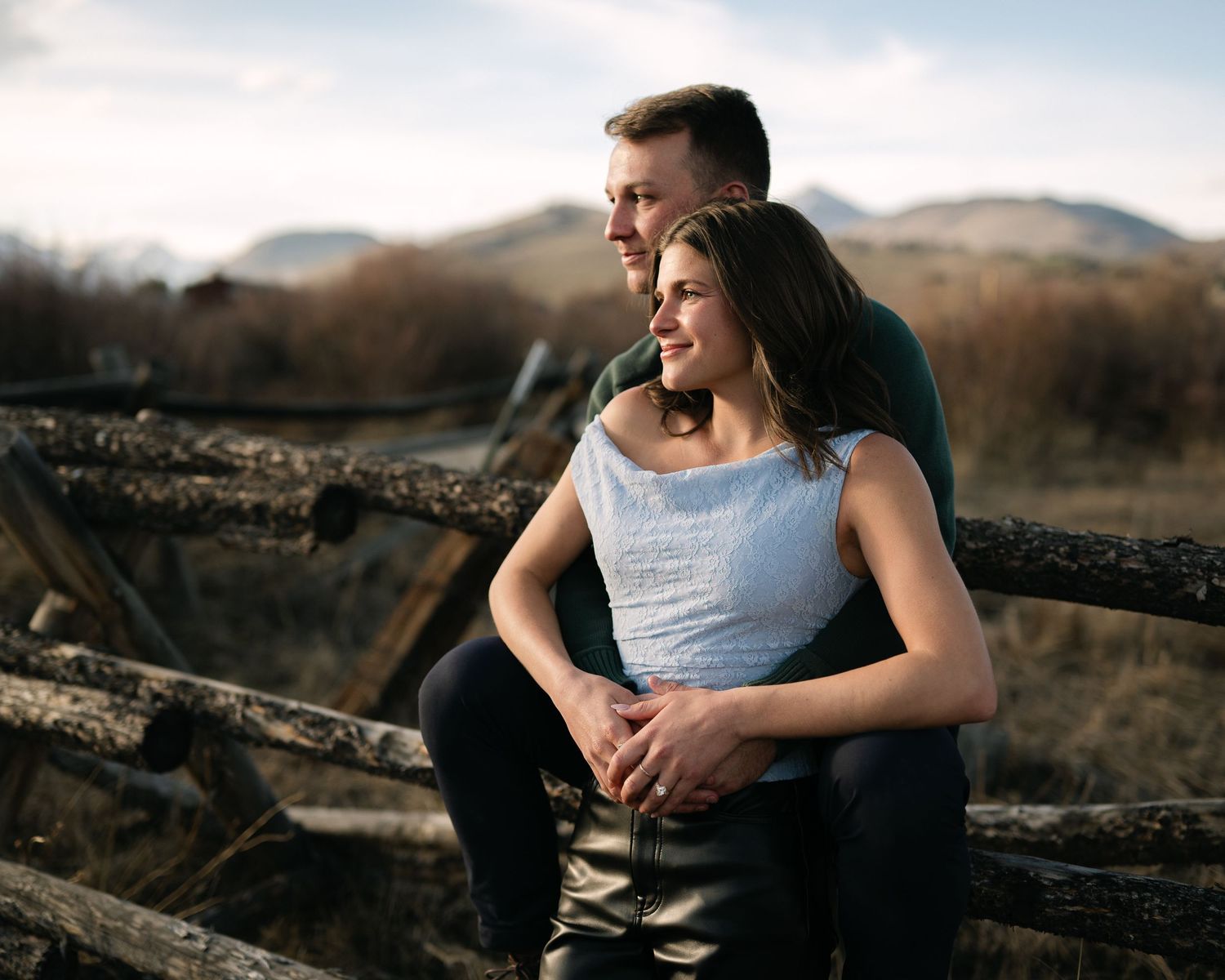 Couple embracing on rustic wooden fence in a scenic mountain field during golden hour.