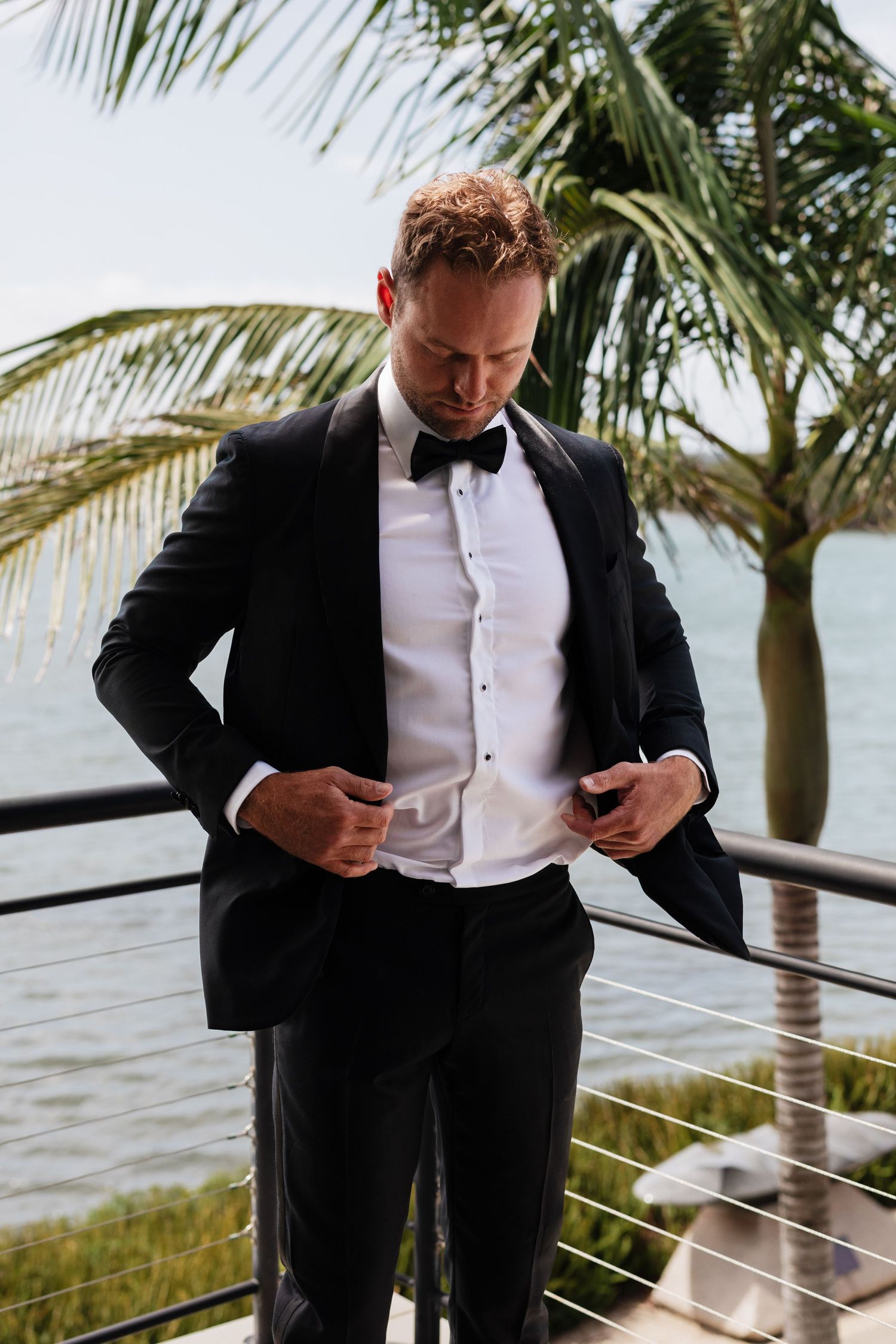 Groom in black tuxedo and bow tie holding a drink, leaning on railing with tropical water view.