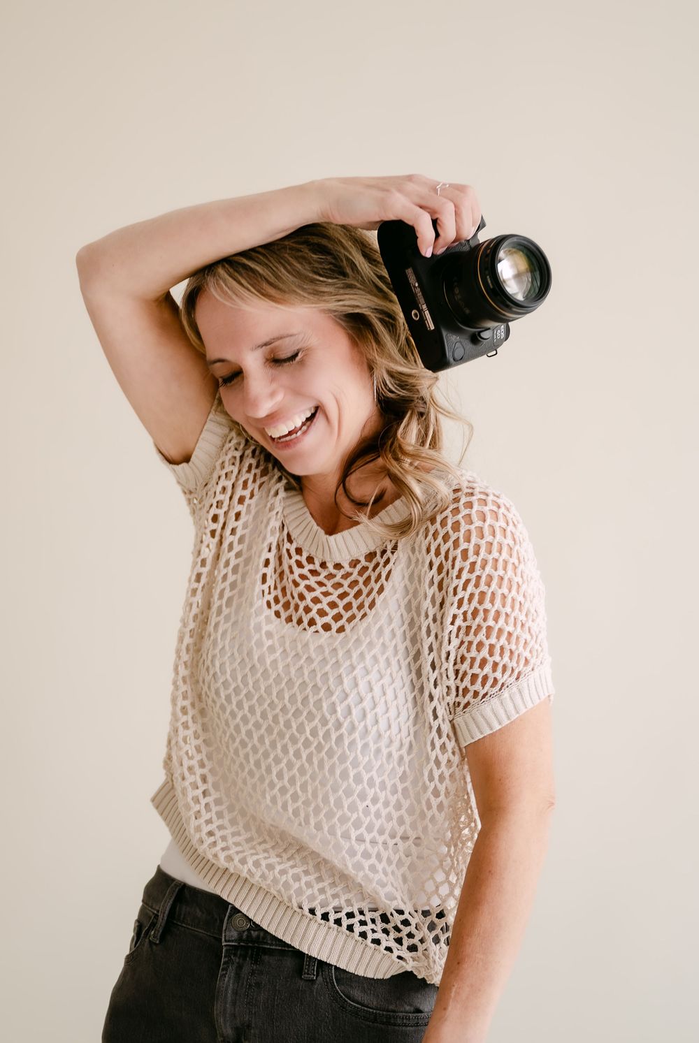 Smiling woman in beige mesh top holding a DSLR camera above her head against a neutral background.