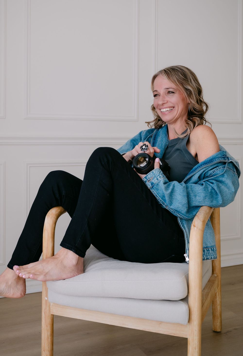 Smiling woman in denim jacket and black pants sitting relaxed in a wooden chair holding a small dark object.