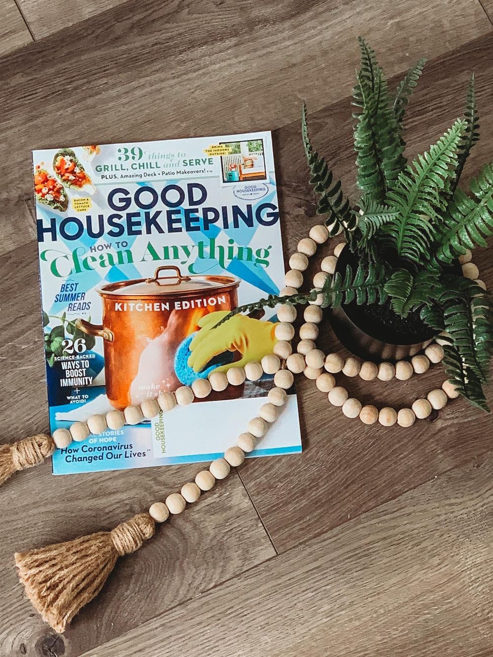 Good Housekeeping magazine with wooden bead garland and potted fern on wood floor.